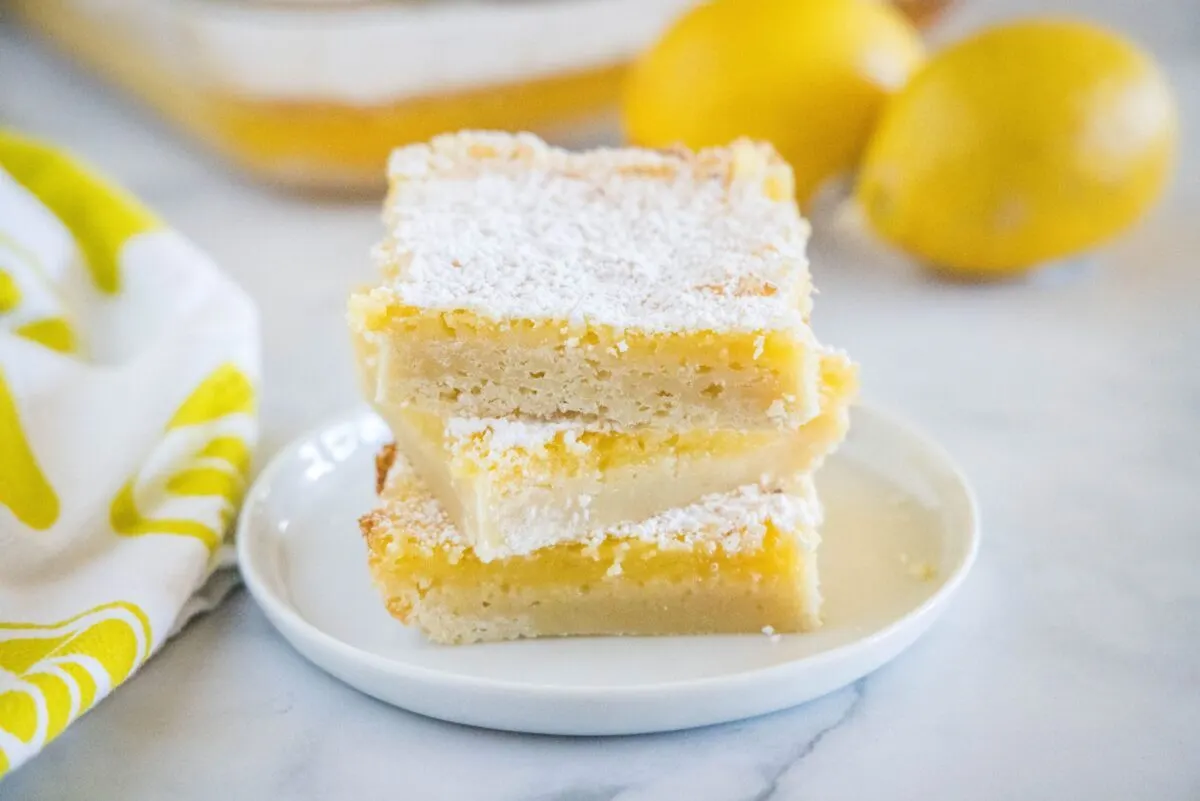 Three lemon bars stacked on each other on a plate, with two lemons behind them