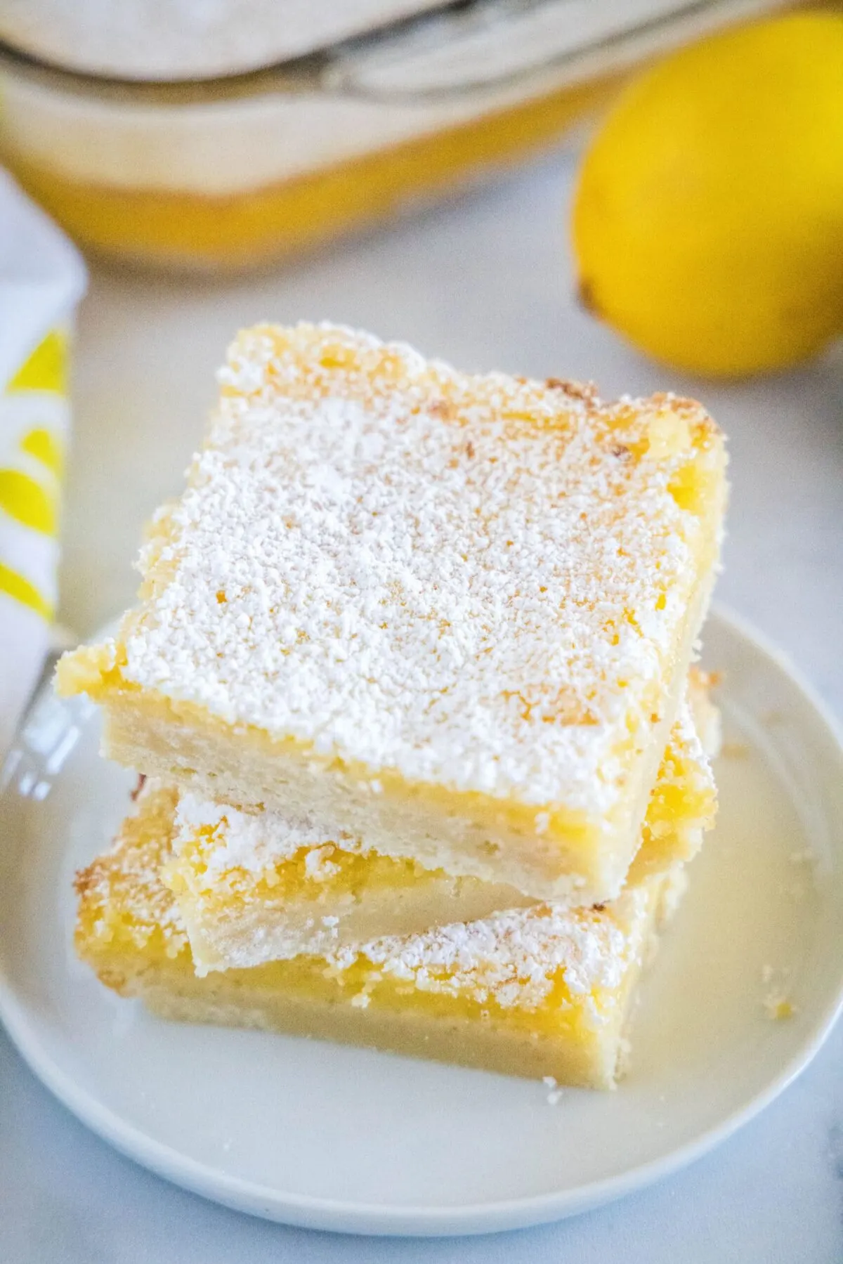 Three square lemon bars stacked on each other on a plate, next to a lemon