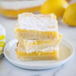 Three square lemon bars stacked on a plate with more bars and lemons behind them