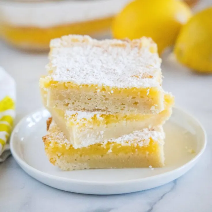 Three square lemon bars stacked on a plate with more bars and lemons behind them