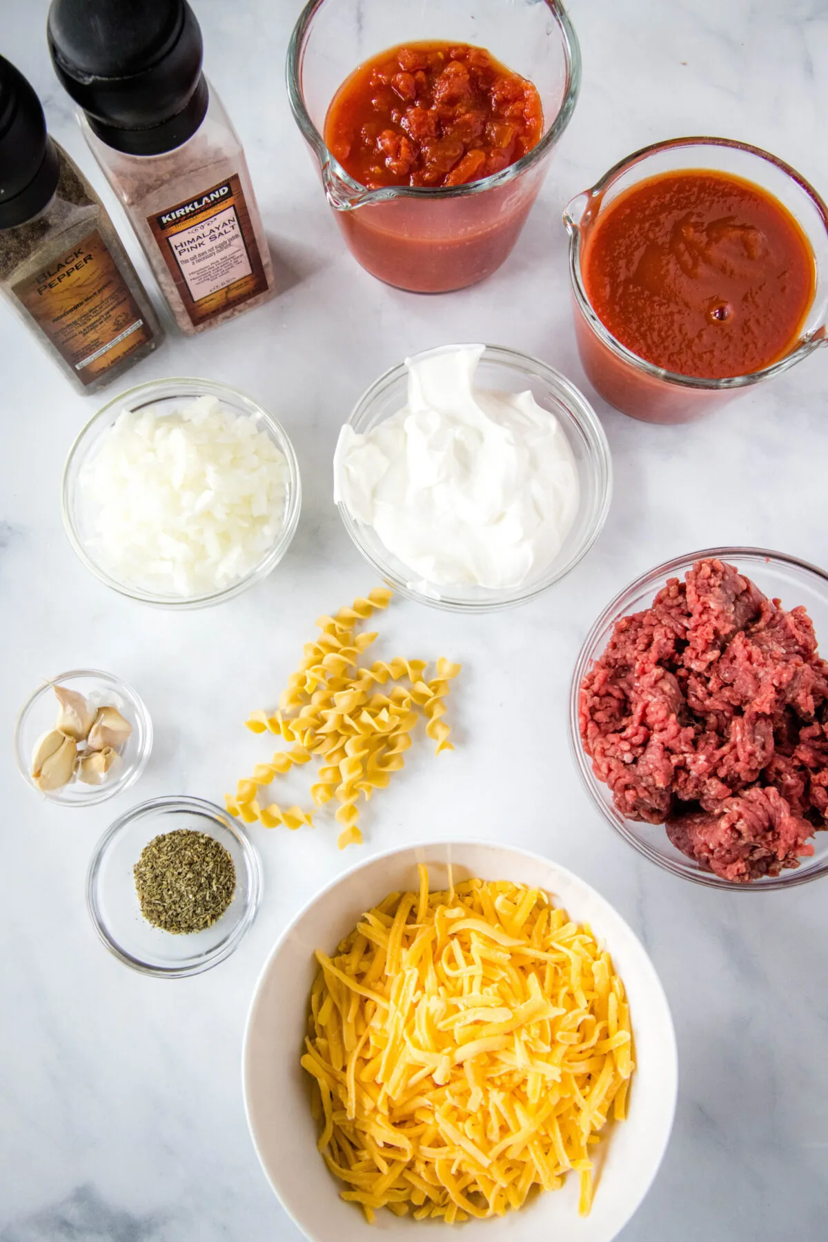 Overhead view of the ingredients needed for beef noodle casserole: a bowl of ground beef, a bowl of shredded cheese, a bowl of sour cream, a bowl of onions, a bowl of pepper, a bowl of garlic, a bowl of Italian seasoning, a jar of tomatoes, a jar of tomato sauce, some scattered egg noodles, and salt and pepper grinders