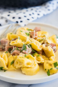 Ham tortellini garnished with chopped parsley and parmesan on a white plate, with a fork in the background.