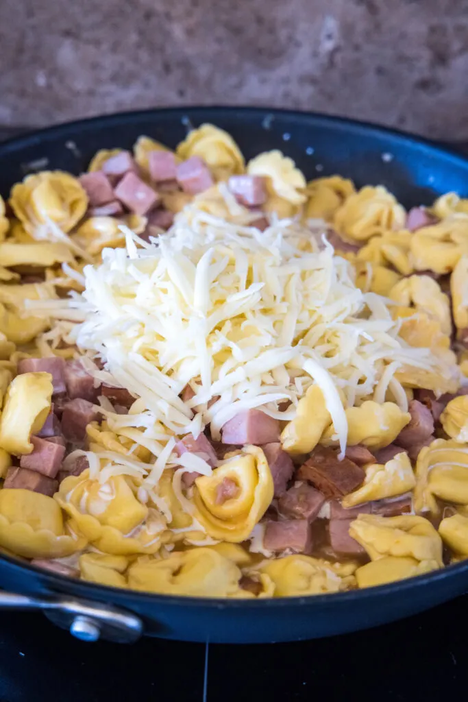 Shredded cheese added to a skillet with ham tortellini.