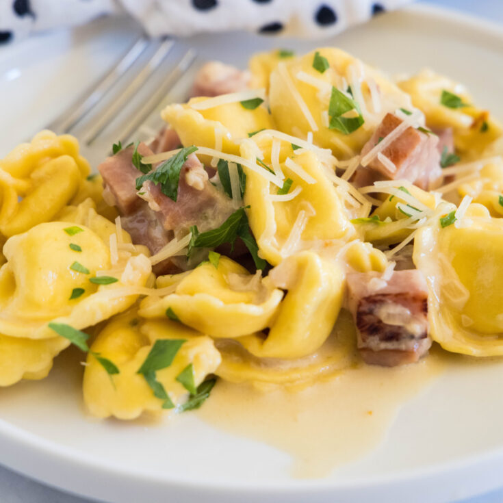 Ham tortellini garnished with chopped parsley and parmesan on a white plate, with a fork in the background.