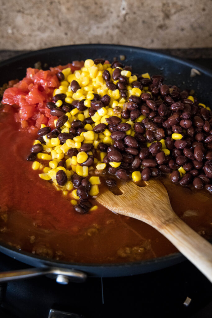 A wooden spatula stirs black beans, corn, and diced tomatoes into a skillet with the ground beef lasagna filling.