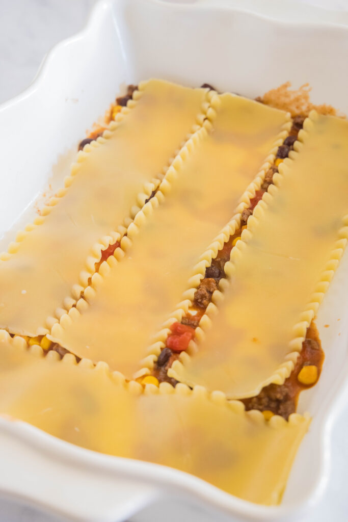 Lasagna noodles layered over Mexican ground beef filling in a white ceramic baking dish.