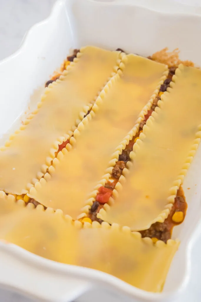 Lasagna noodles layered over Mexican ground beef filling in a white ceramic baking dish.