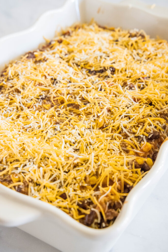 Assembled Mexican lasagna topped with shredded cheese in a white ceramic baking dish.