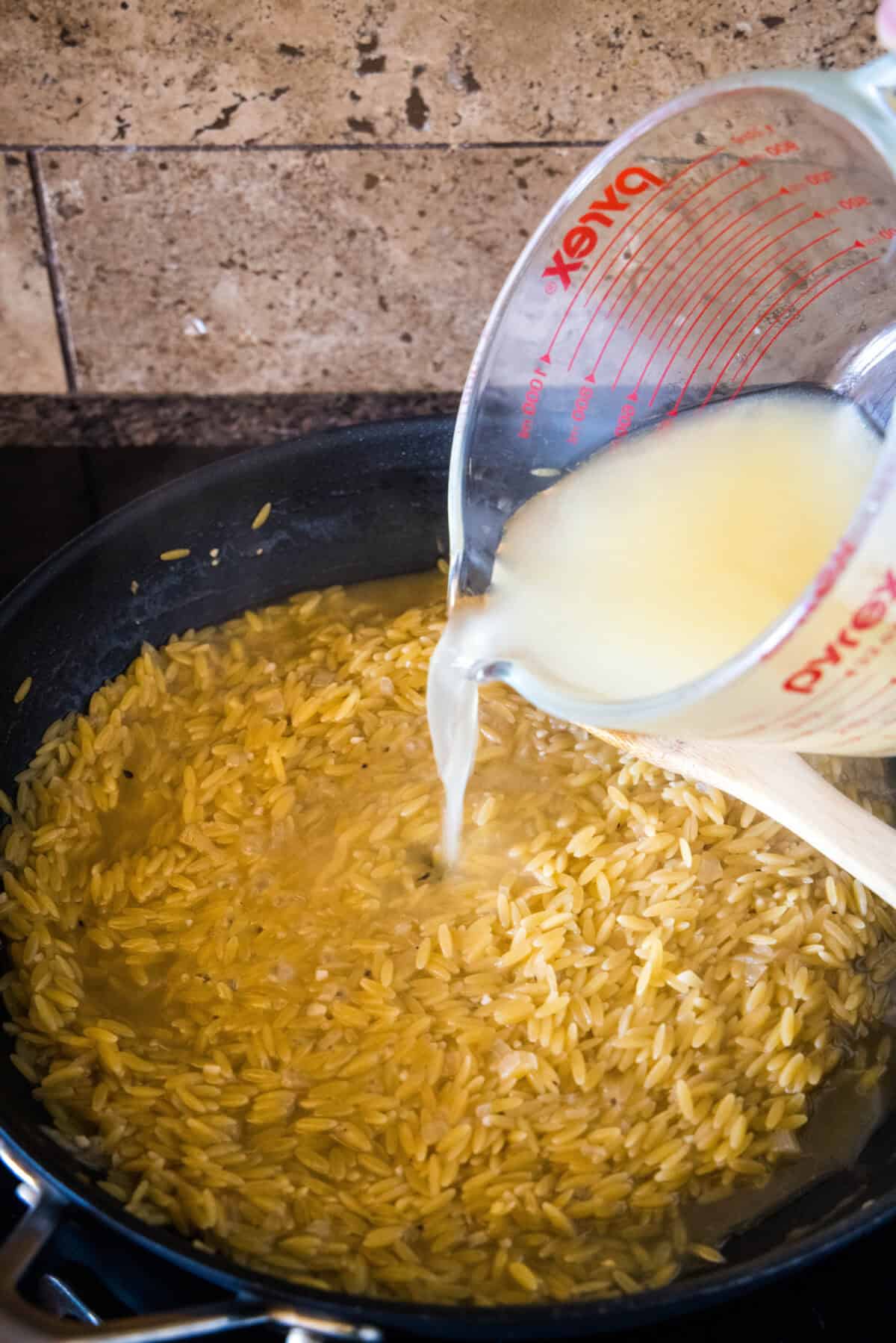 Broth being poured into a skillet with dried orzo.