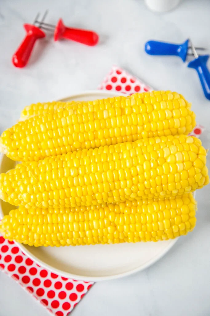 fresh corn on the cob staked on a white plate with red and white napkin 