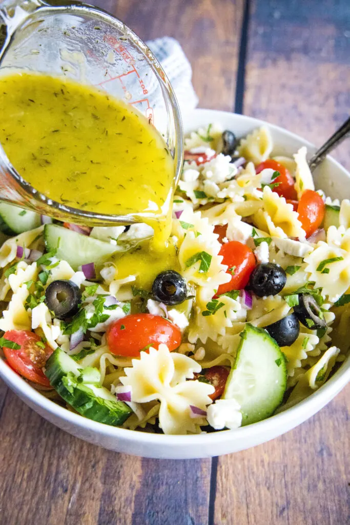 pouring greek dressing over a bowl of pasta salad