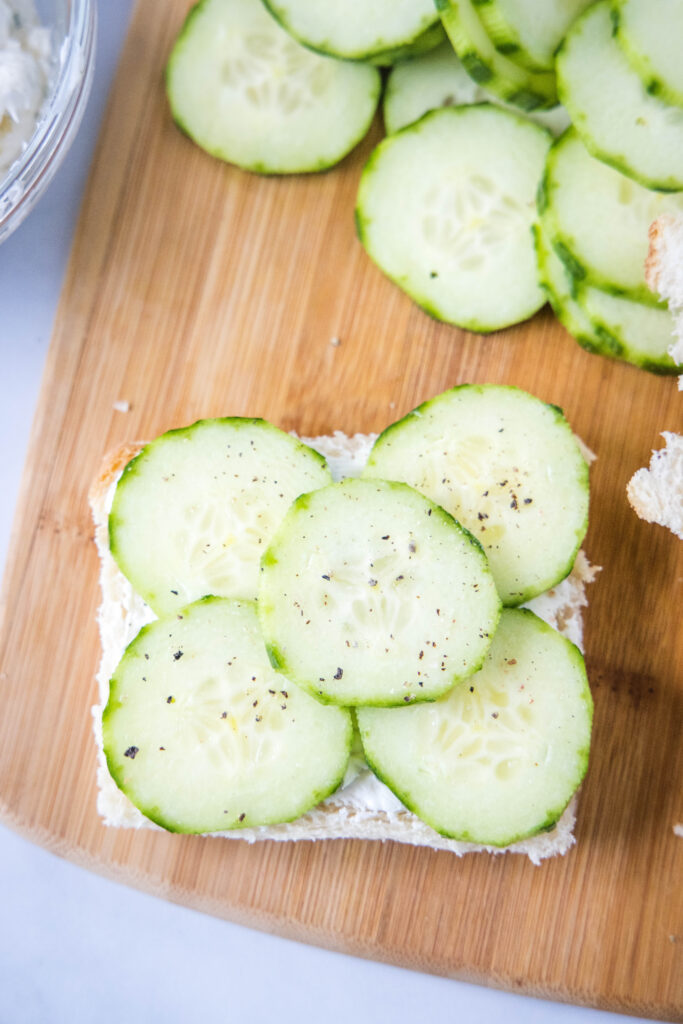 placing cucumbers on a slice of bread with cream cheese spread on it