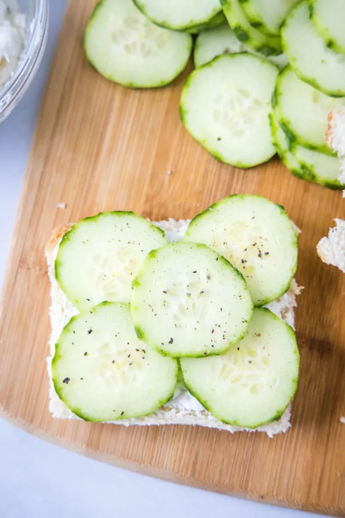 placing cucumbers on a slice of bread with cream cheese spread on it