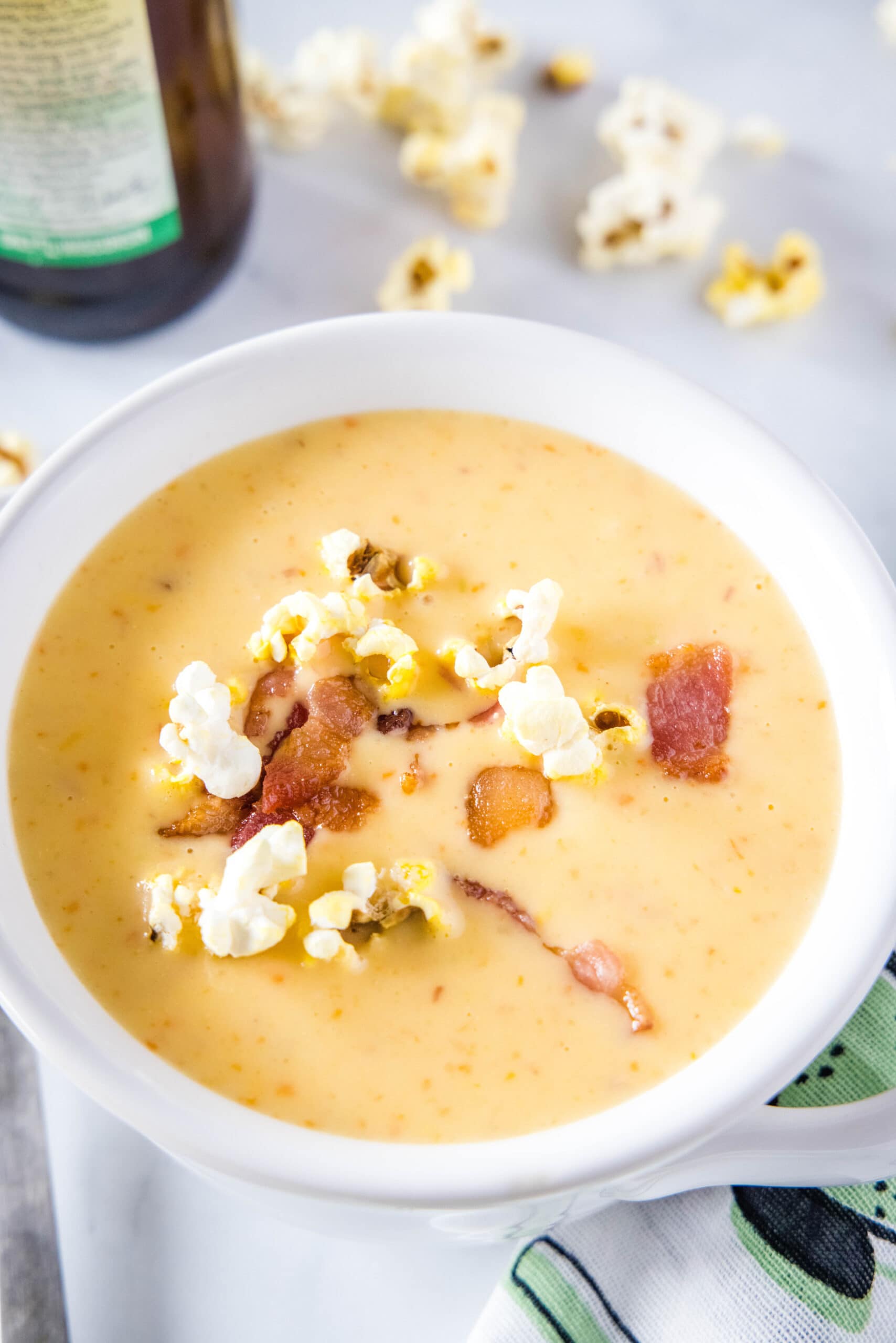 A bowl of cheese soup topped with popcorn and bacon, with a bottle of beer and some popcorn in the background