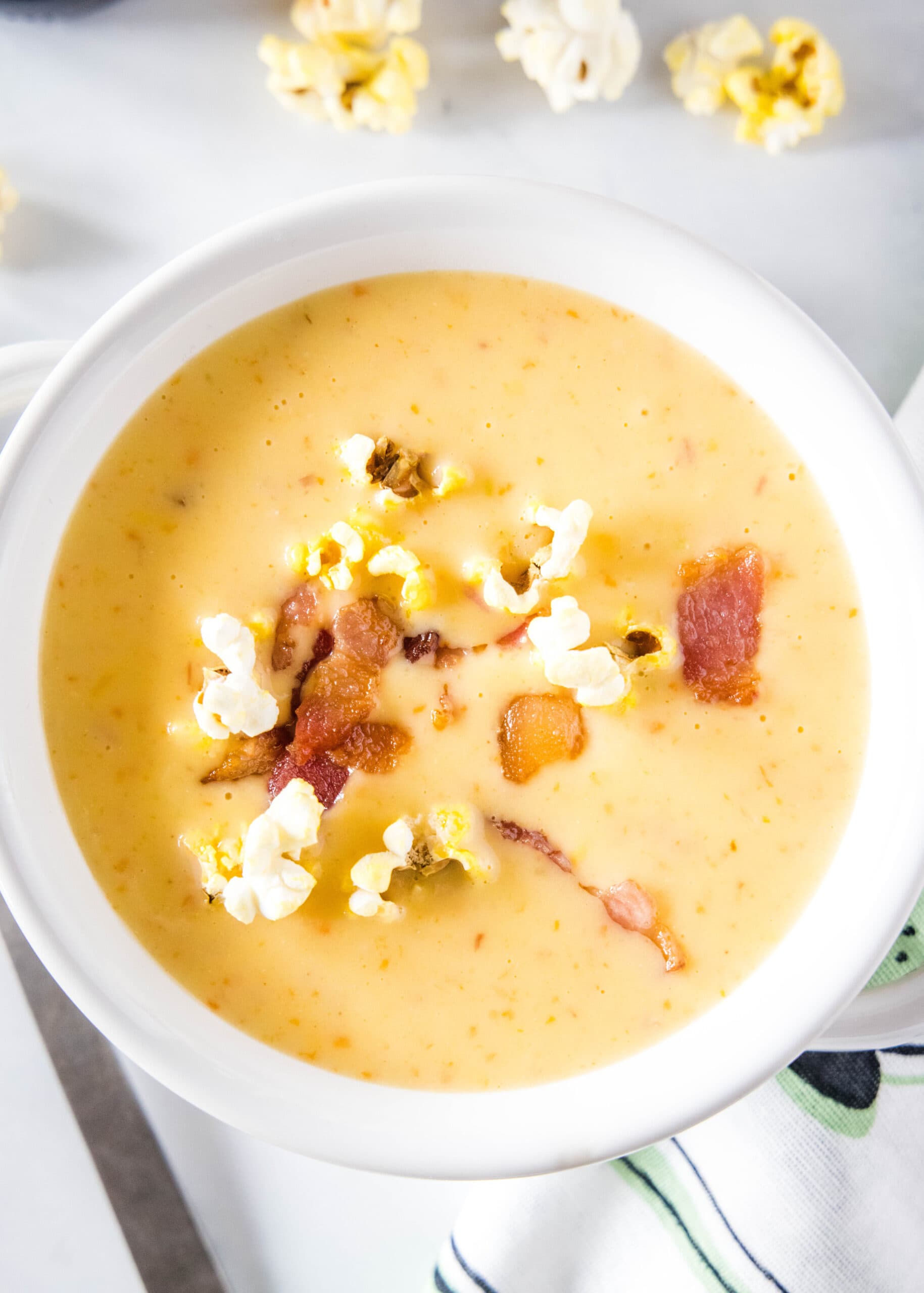 Overhead view of a bowl of cheese soup topped with popcorn and bacon, with some popcorn on the side