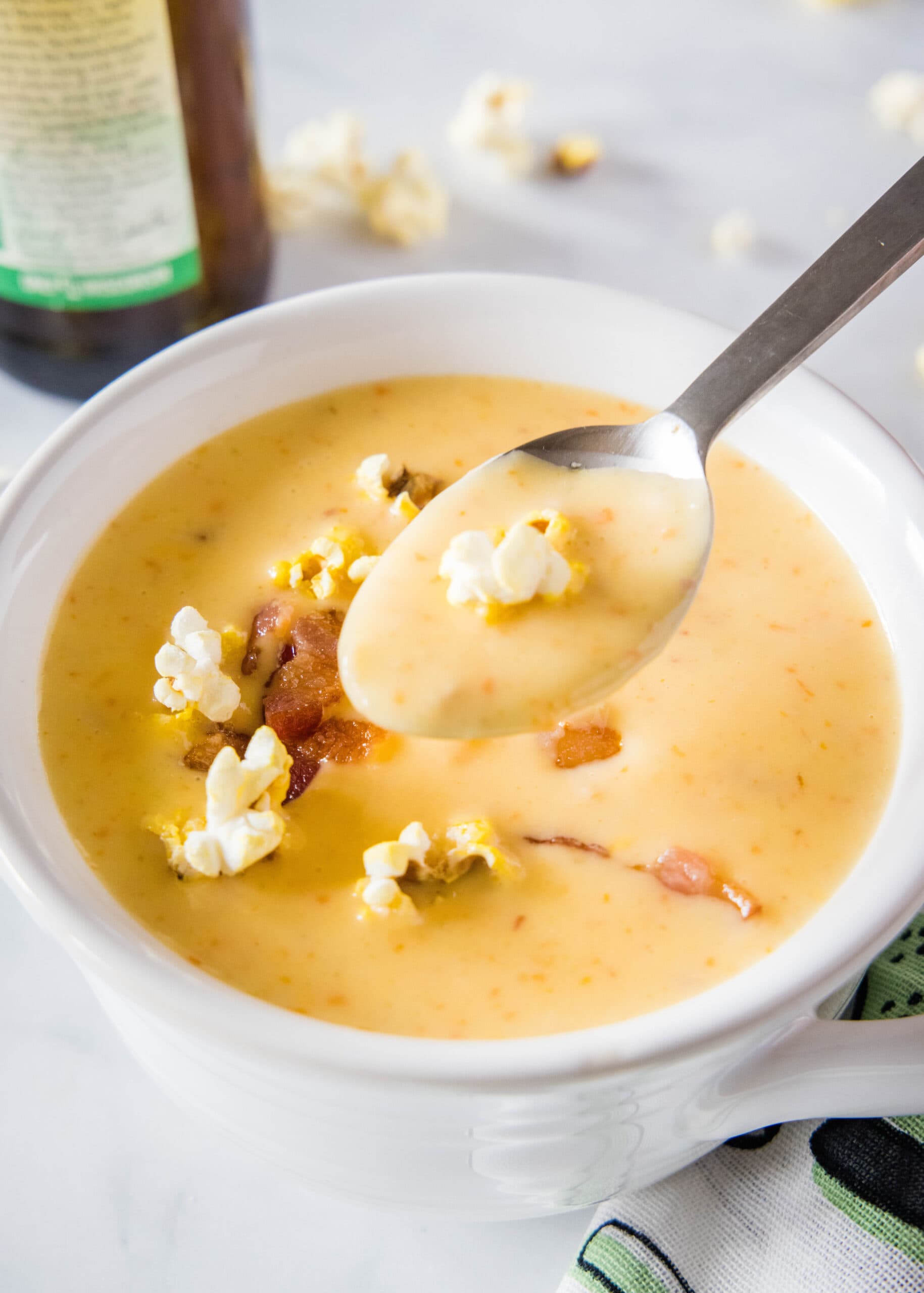 A bowl of cheese soup topped with bacon and popcorn, with a spoon taking a spoonful out, and a bottle of beer in the background
