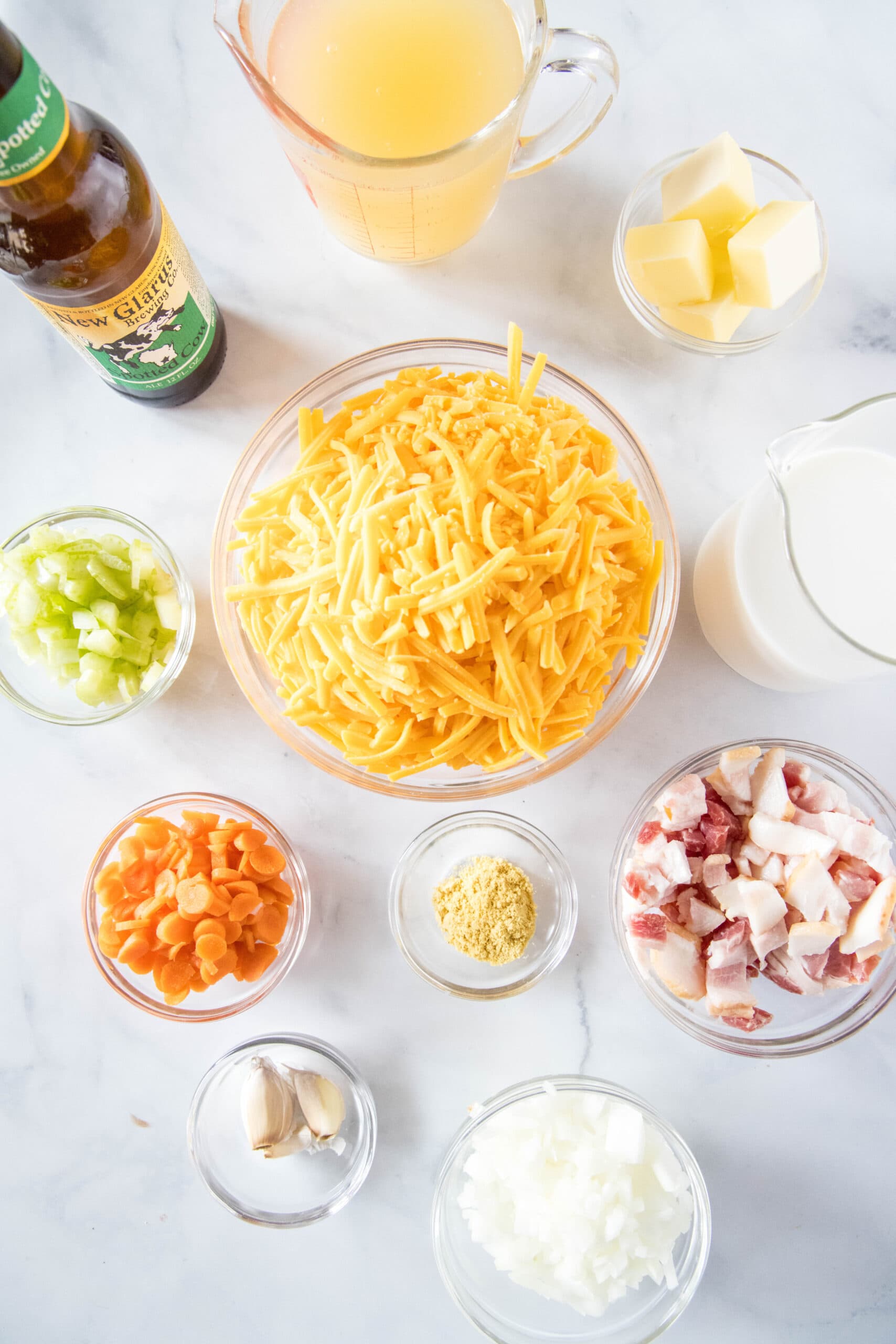 Overhead view of the ingredients needed for beer cheese soup: a bottle of beer, a bowl of butter, a bowl of grated cheddar cheese, a pitcher of milk, a bowl of diced celery, a bowl of diced carrots, a bowl of dried mustard, a bowl of diced onions, a bowl of garlic cloves, and a bowl of chopped bacon