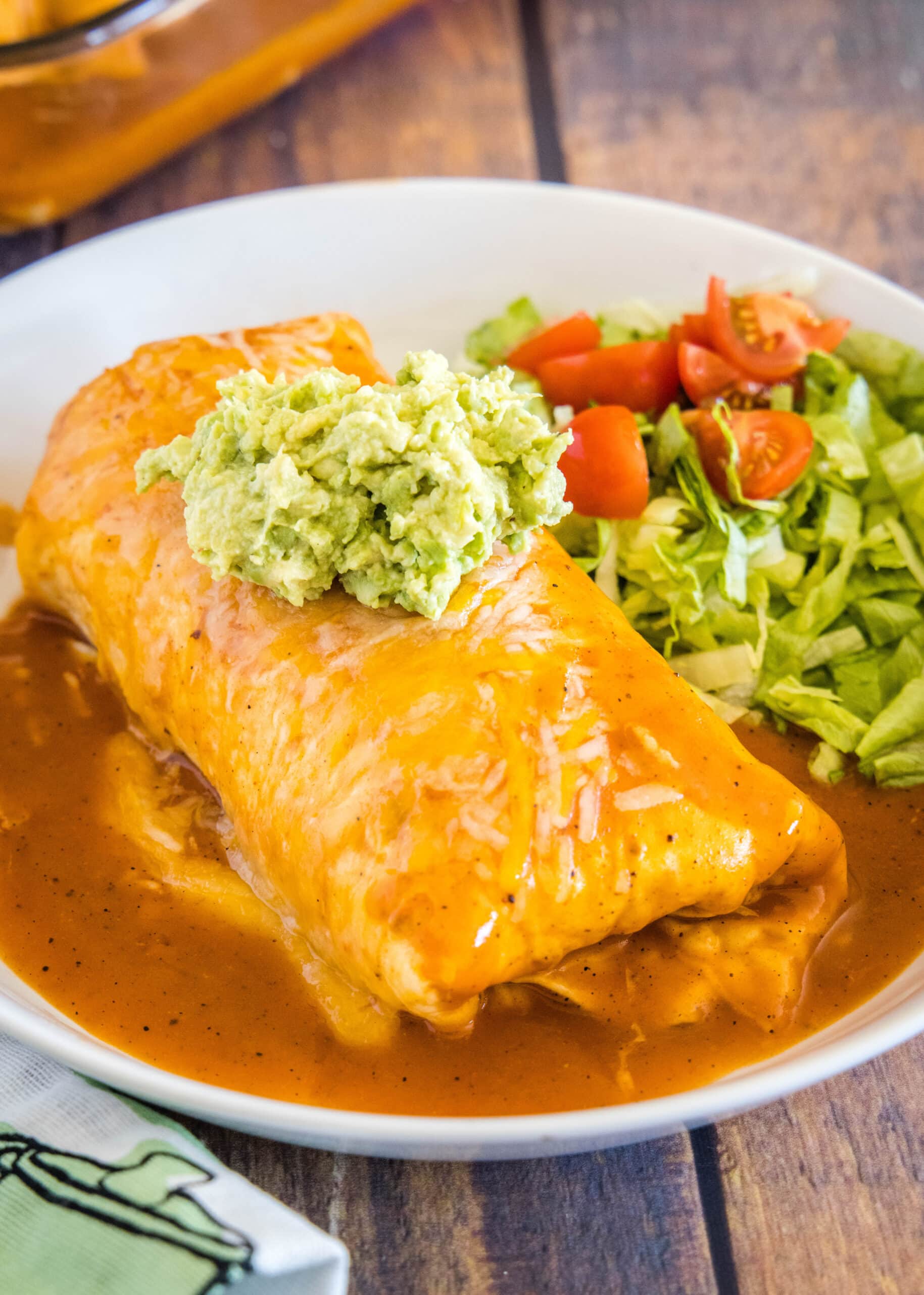 A wet burrito on a white plate, topped with guacamole, next to lettuce and tomatoes