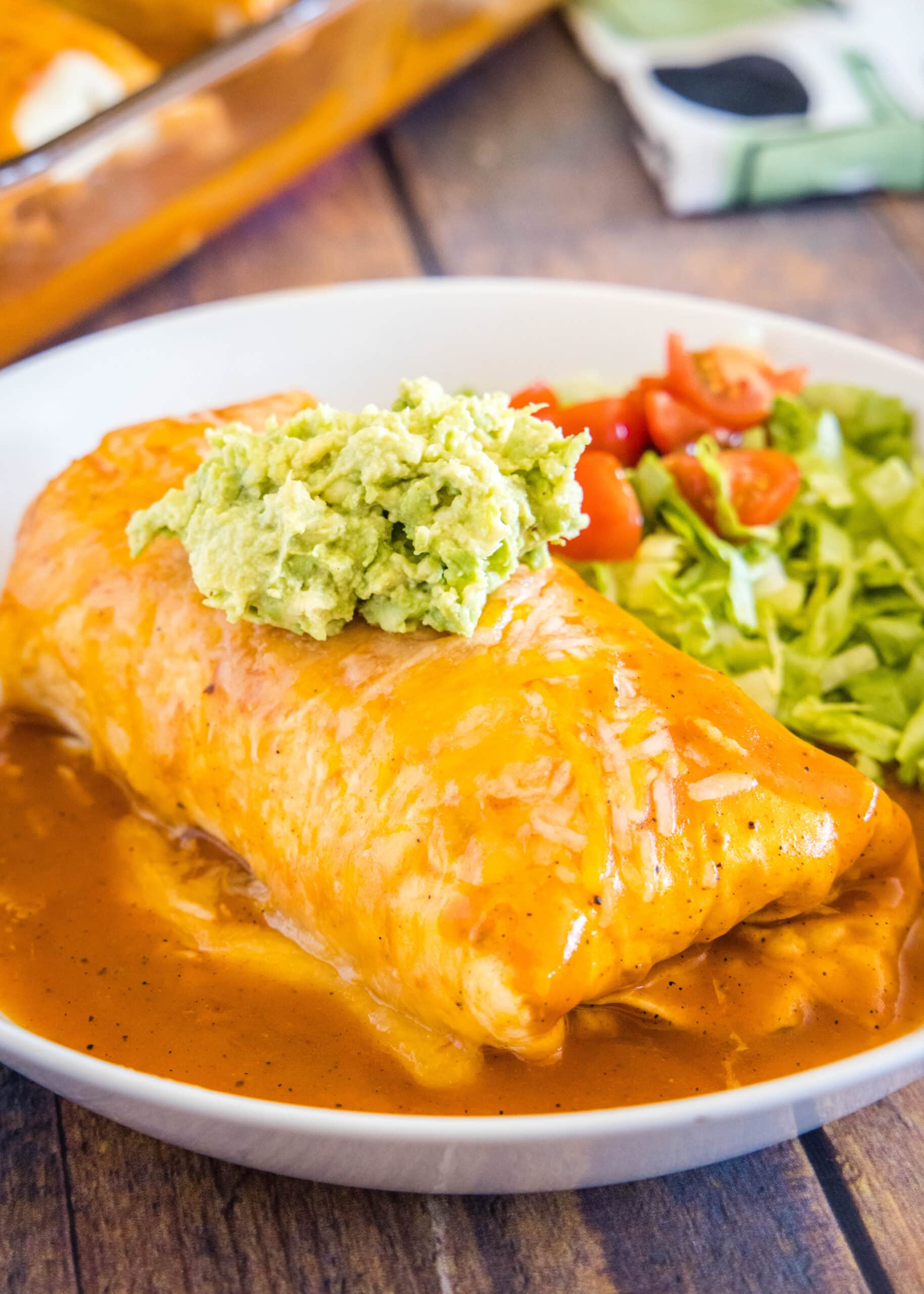 A wet burrito on a plate topped with guacamole, with a salad of lettuce and tomatoes in the background.