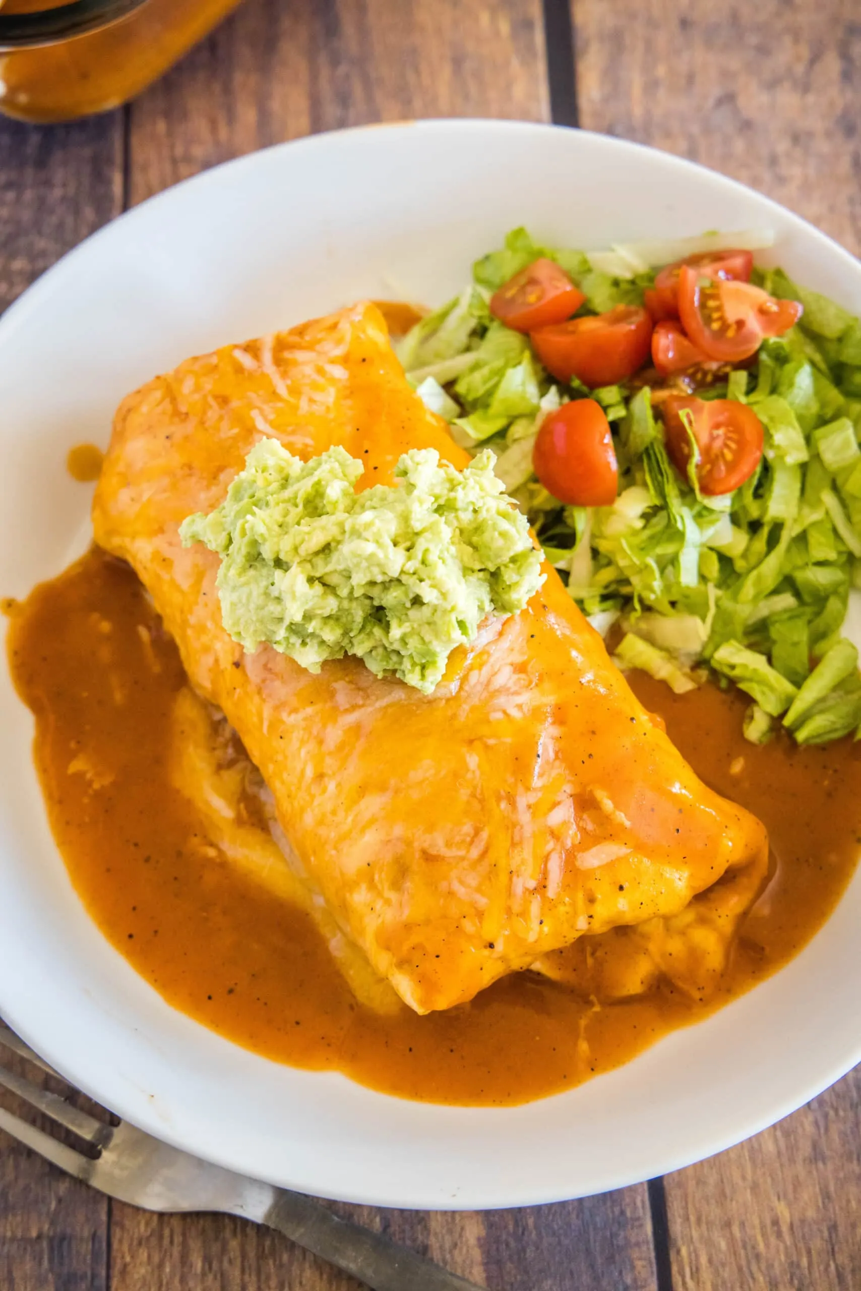 Overhead view of a wet burrito topped with guacamole, on a plate with lettuce and tomatoes, next to a fork