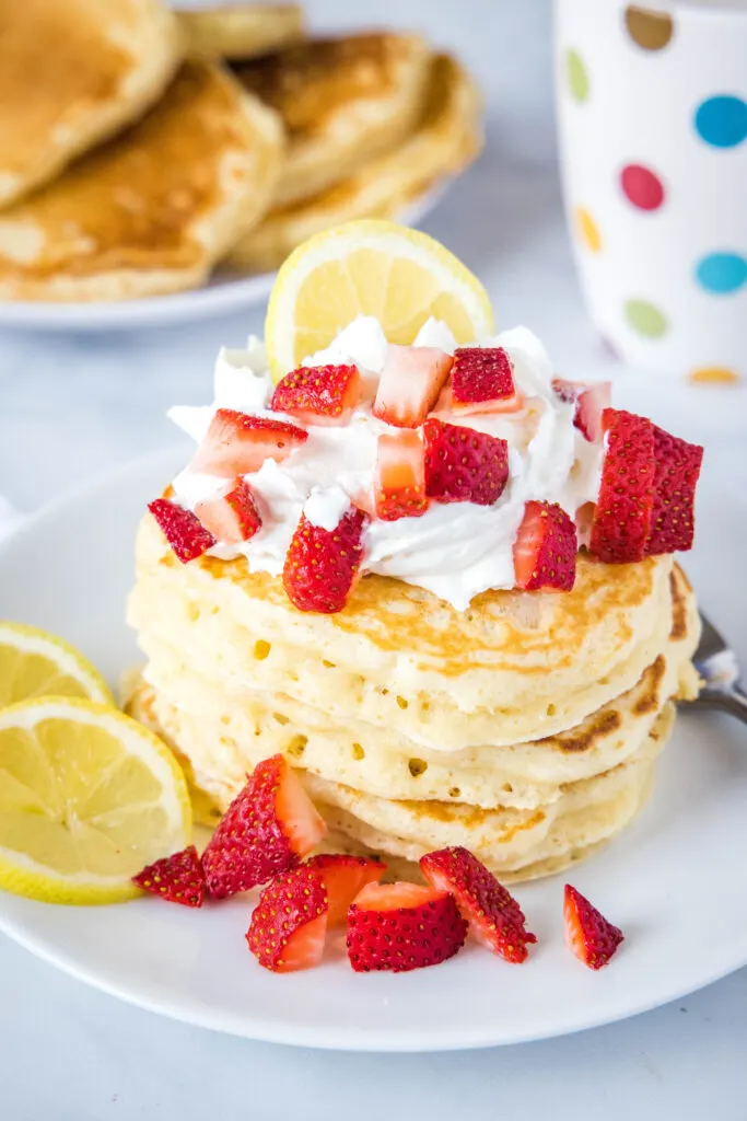 lemon pancakes stacked on a white plate with whipped cream and strawberries