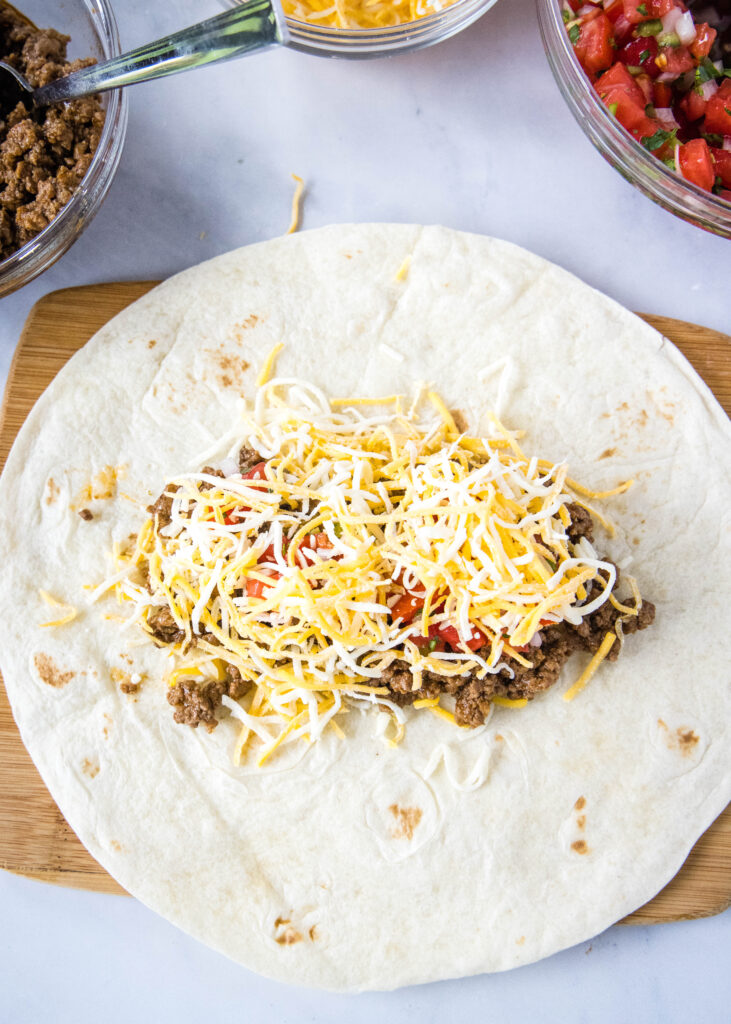 adding cheese to tortilla over tomatoes and ground beef