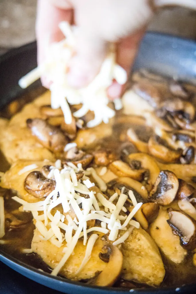 sprinkling cheese over chicken and mushrooms in a skillet