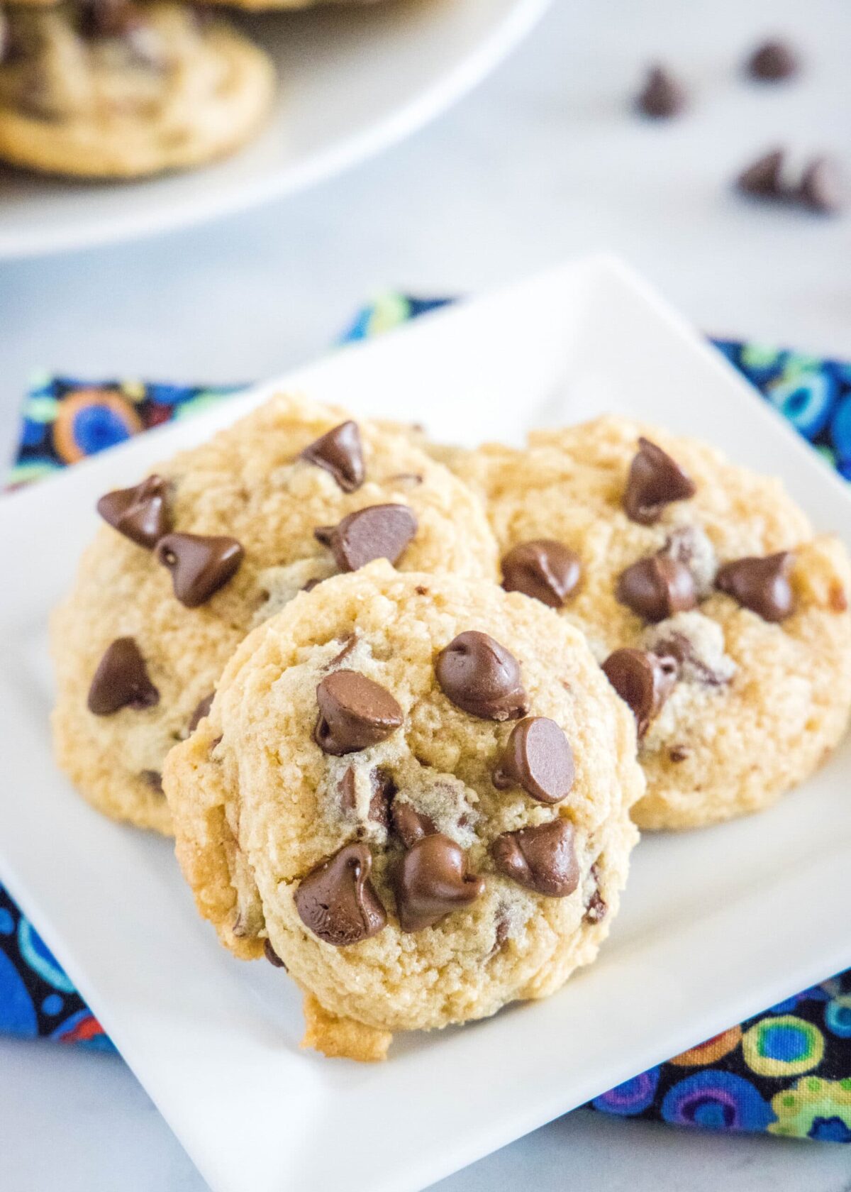 Close up of three chocolate chip cookies on a white plate on top of a kitchen towel, with cookies and chocolate chips in the background