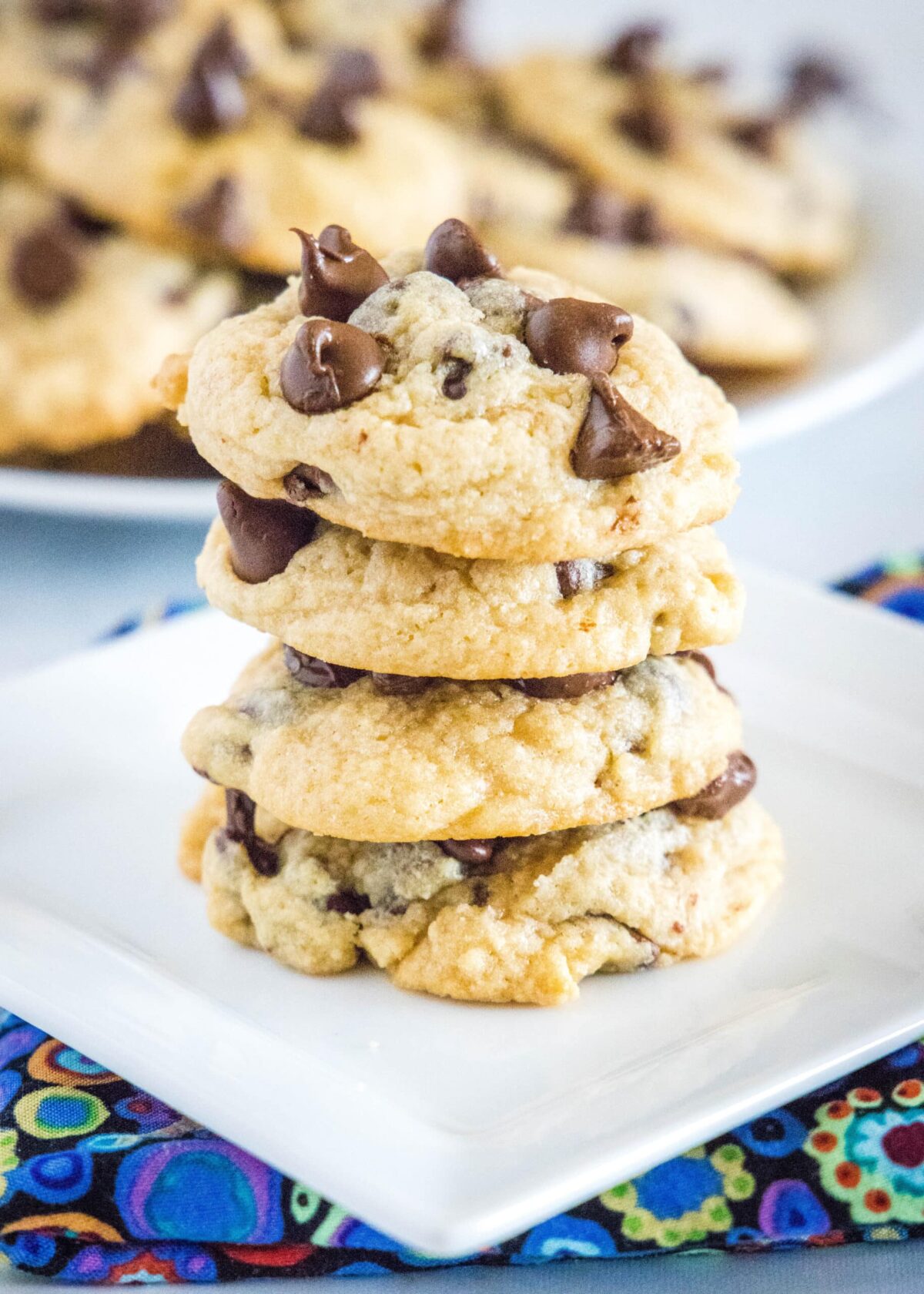 Four chocolate chip cookies stacked on a plate, with more cookies in the background