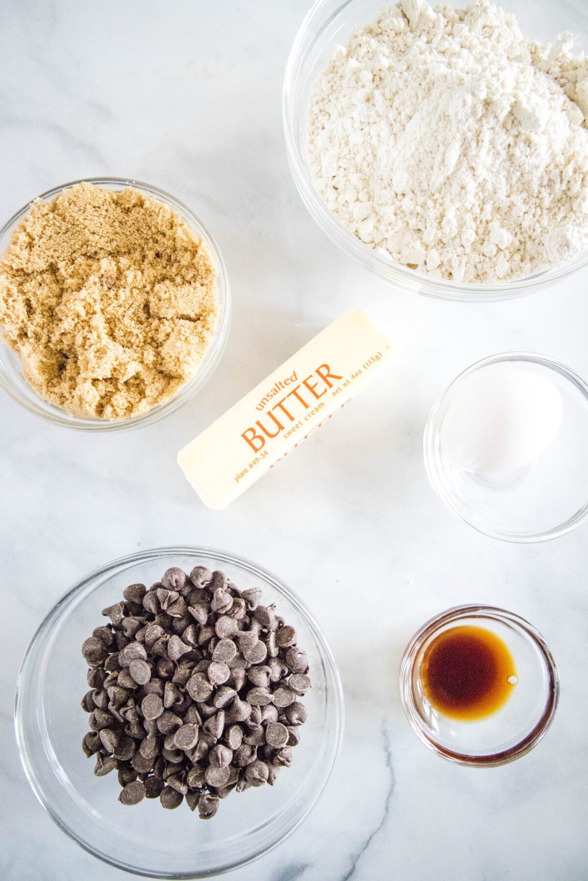 Overhead view of the ingredients needed for Bisquick cookies: A bowl of Bisquick, a bowl of brown sugar, a bowl of chocolate chips, a stick of butter, a ramekin of vanilla, and a bowl with an egg