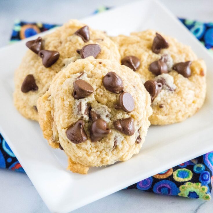 Three chocolate chip cookies on a white square plate, on top of a kitchen towel