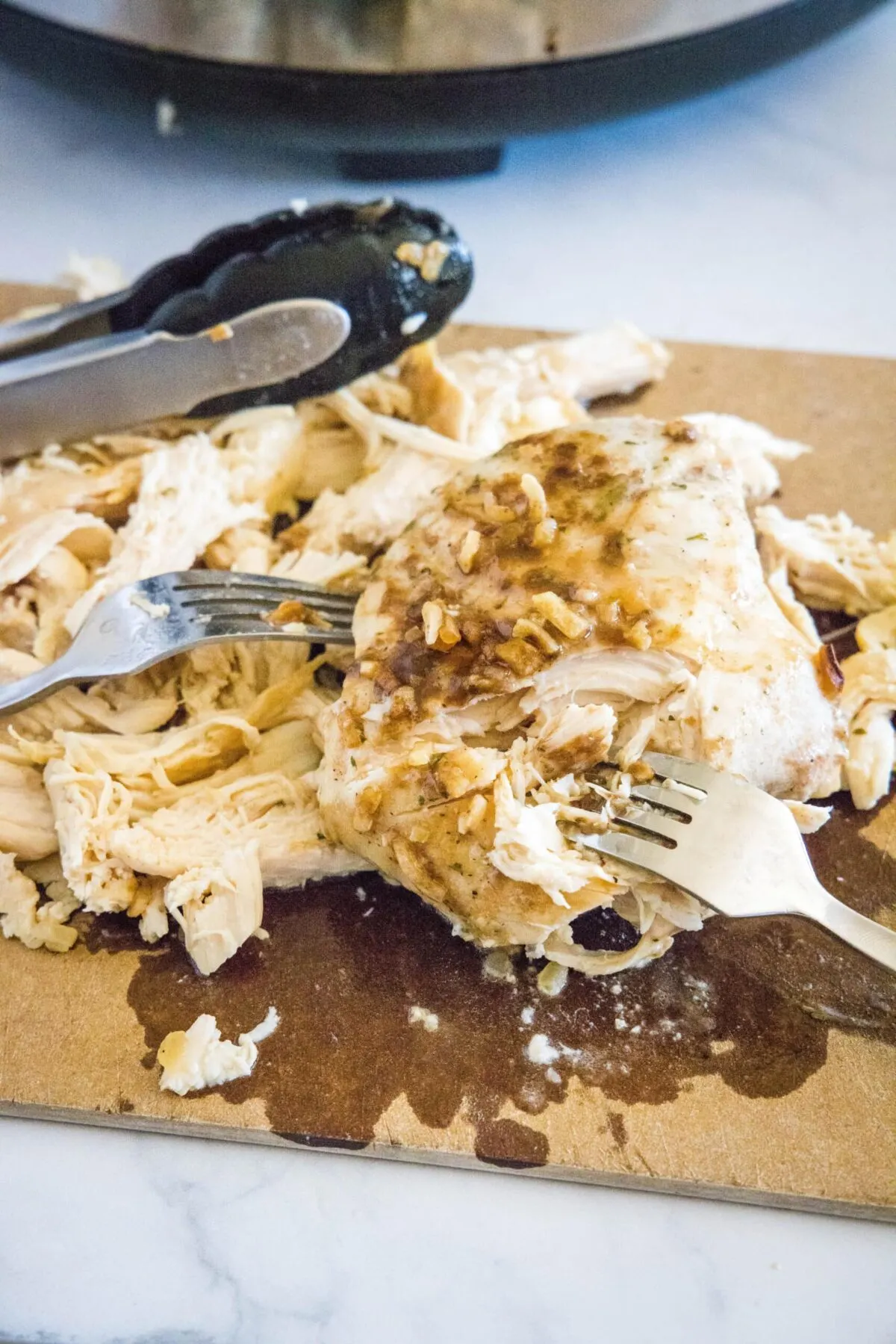 Cooked chicken breasts on a cutting board with a pair of tongs, with two forks shredding them