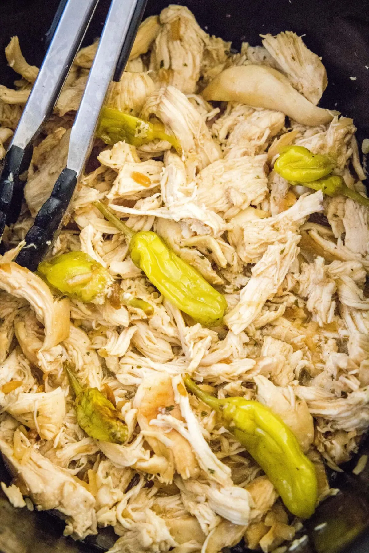 Close up of shredded chicken and pepperoncini peppers in a slow cooker with tongs
