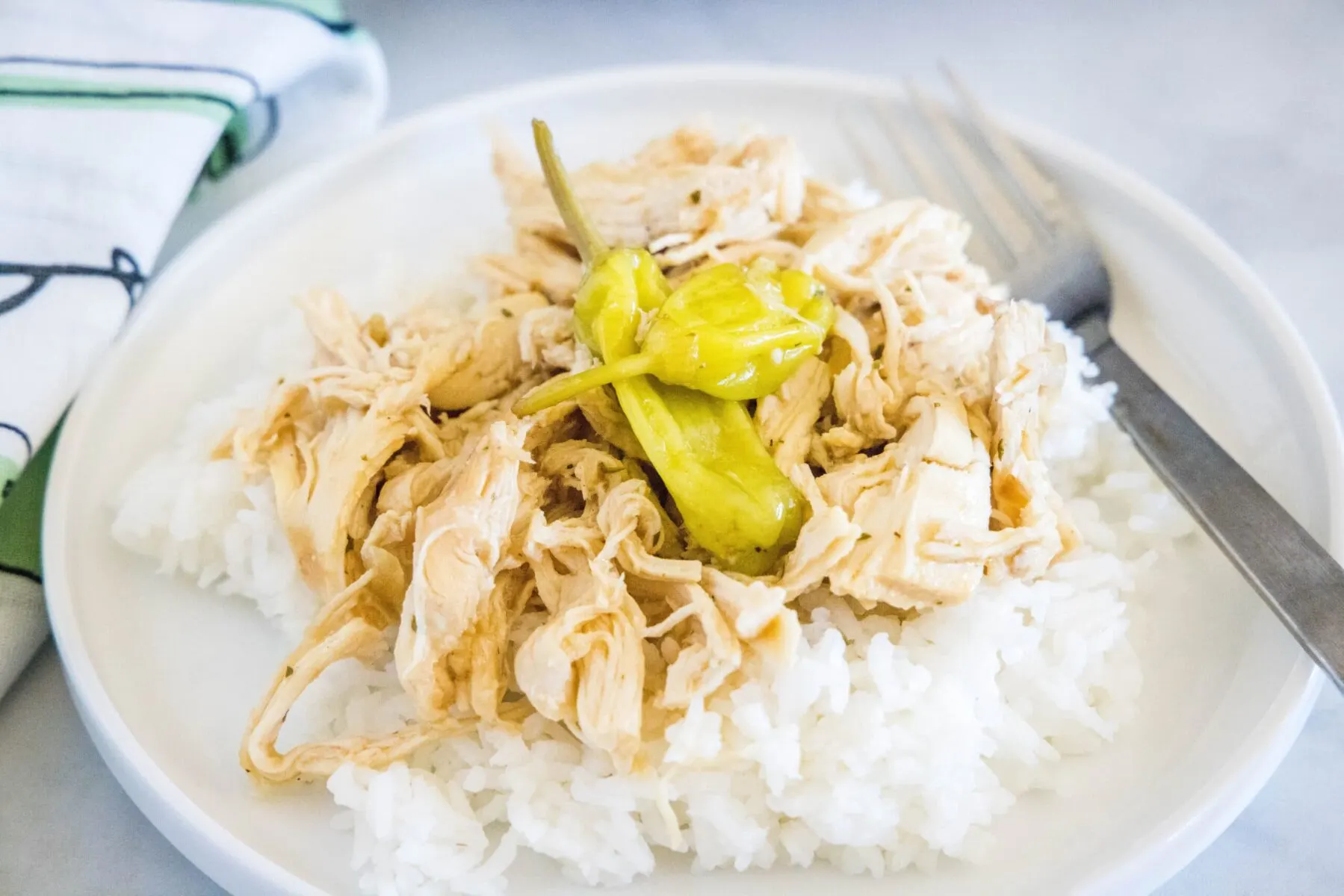A plate with a fork and some rice, topped with shredded chicken and pepperoncini peppers, next to a kitchen towel