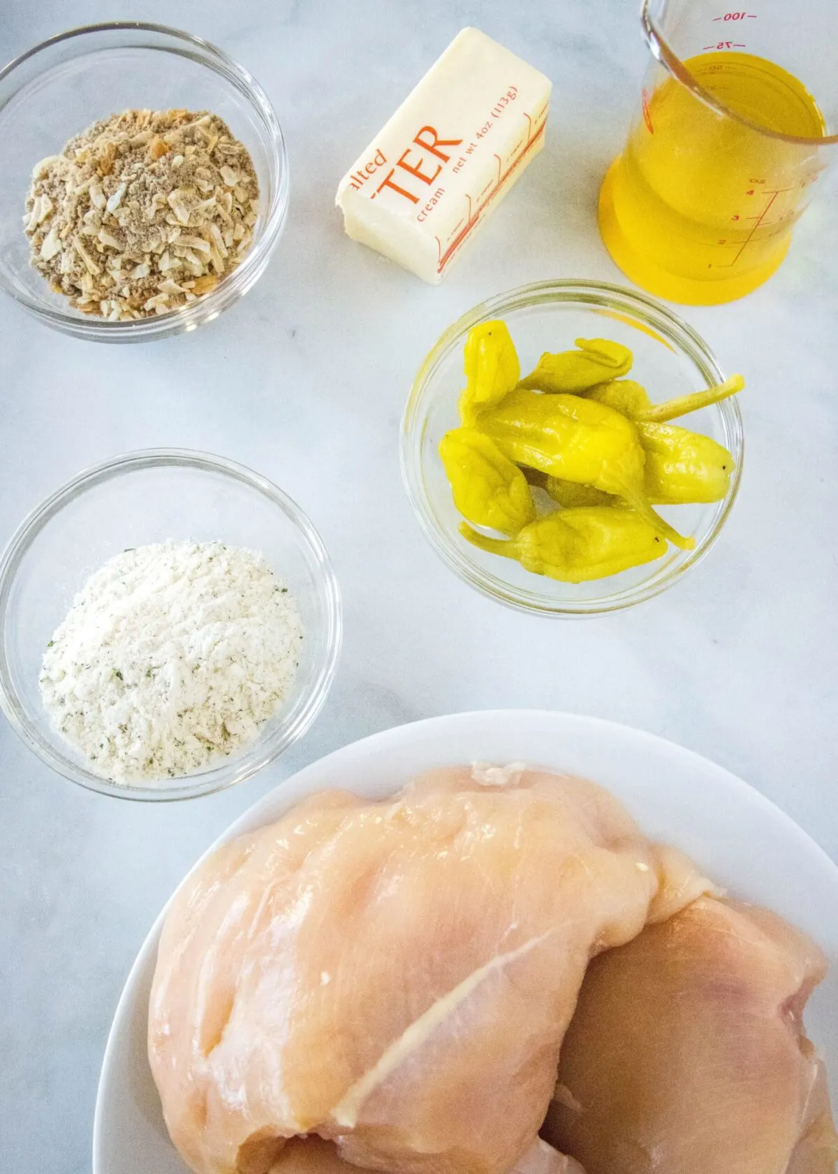 Overhead view of the ingredients needed for Mississippi chicken: a plate of raw chicken breasts, a bowl of ranch dressing mix, a bowl of onion soup mix, a bowl of pepperoncini peppers, a glass of pepperoncini juice, and half a stick of butter