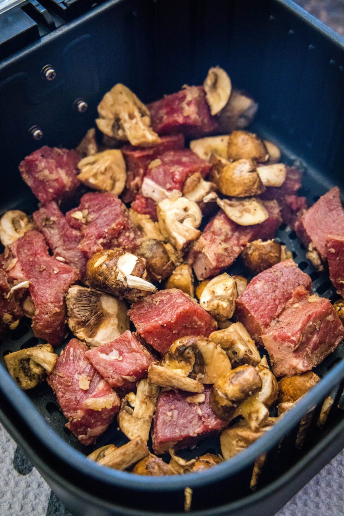 Seasoned, uncooked pieces of steak and mushrooms in an air fryer basket