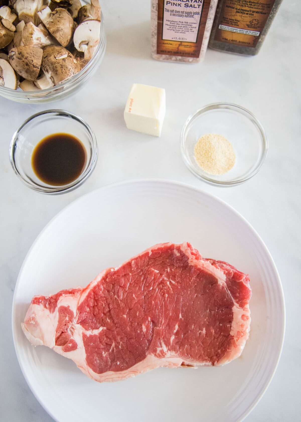 Overhead view of the ingredients needed for air fryer steak: A raw steak on a plate, a bowl of mushrooms, a knob of butter, a bowl of garlic powder, a bowl of Worcestershire sauce, a bottle of salt and a bottle of pepper