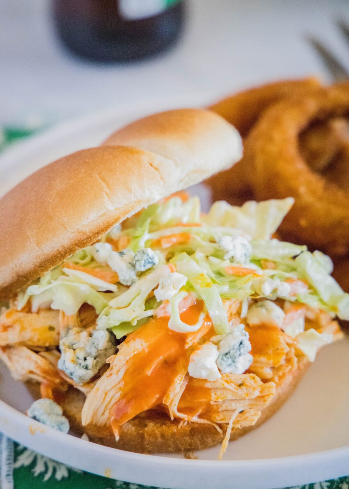A chicken sandwich topped with coleslaw and blue cheese, on a plate, with onion rings in the background