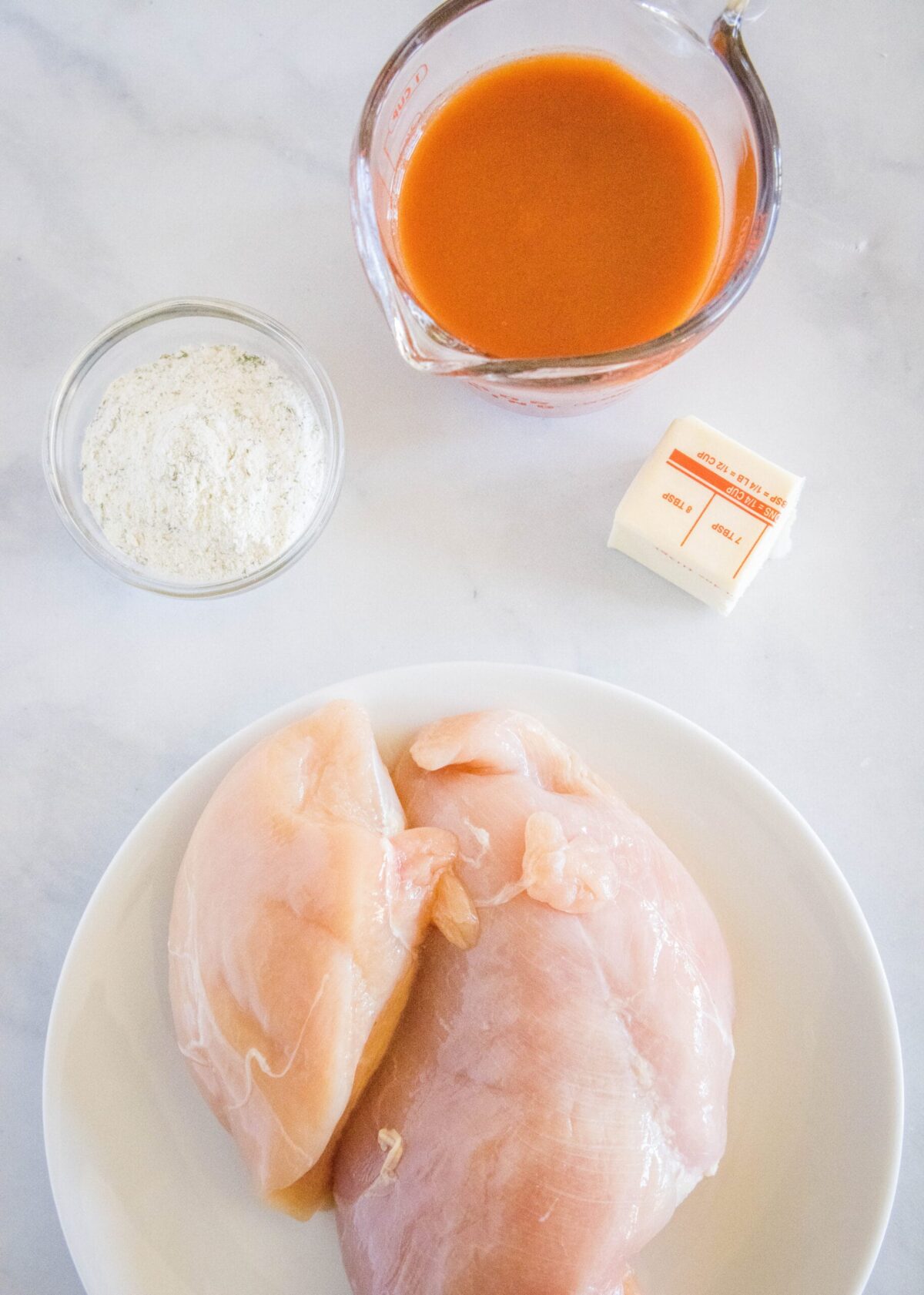 Overhead view of a knob of butter, a plate of raw chicken breasts, a ramekin of ranch dressing mix, and a pyrex of buffalo sauce