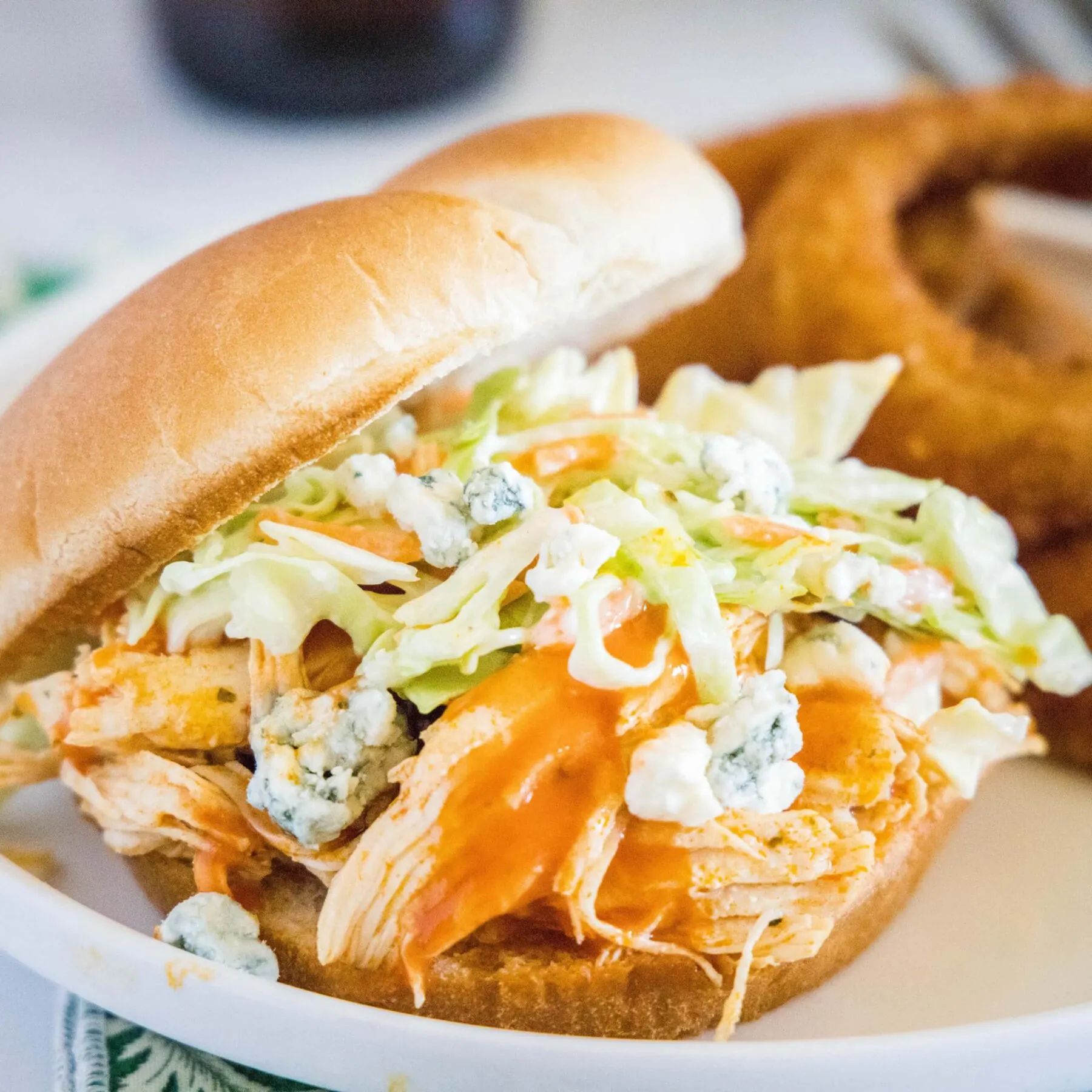 A buffalo chicken sandwich on a plate, topped with coleslaw and blue cheese, with onion rings in the background