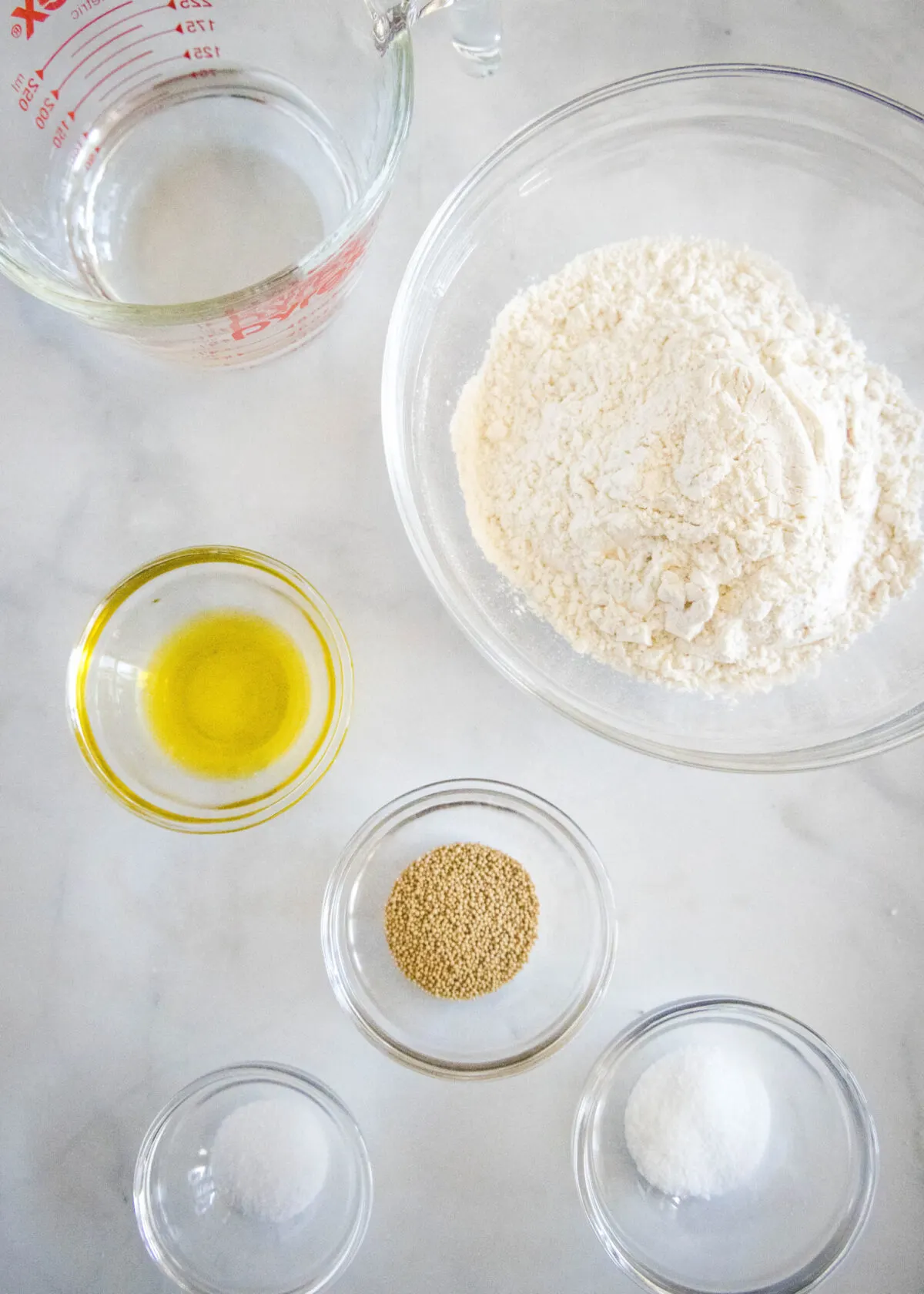 Overhead view of the ingredients needed for pizza dough: a bowl of flour, a bowl of yeast, a bowl of salt, a bowl of olive oil, a glass of water, and a pyrex of water