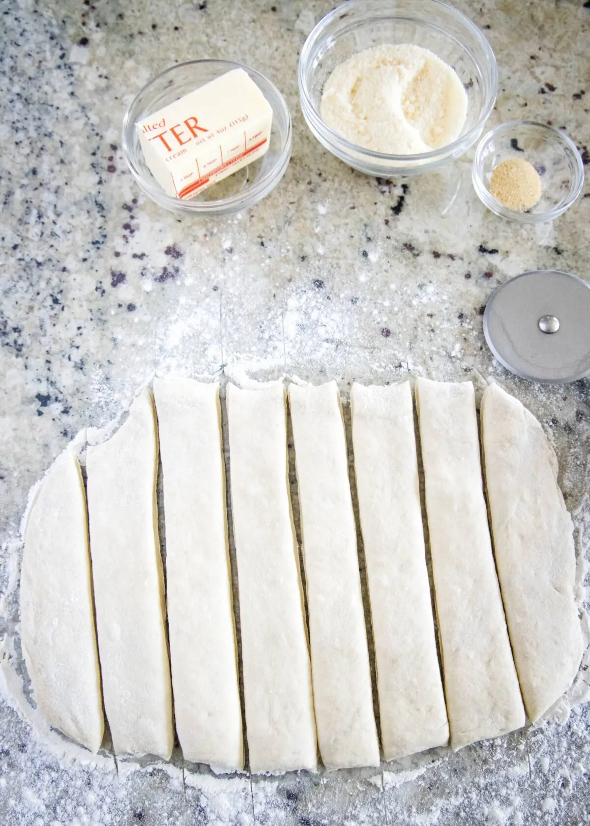 Pizza dough cut into eight strips, next to a pizza cutter, a bowl of parmesan cheese, a bowl of garlic powder, and half a stick of butter
