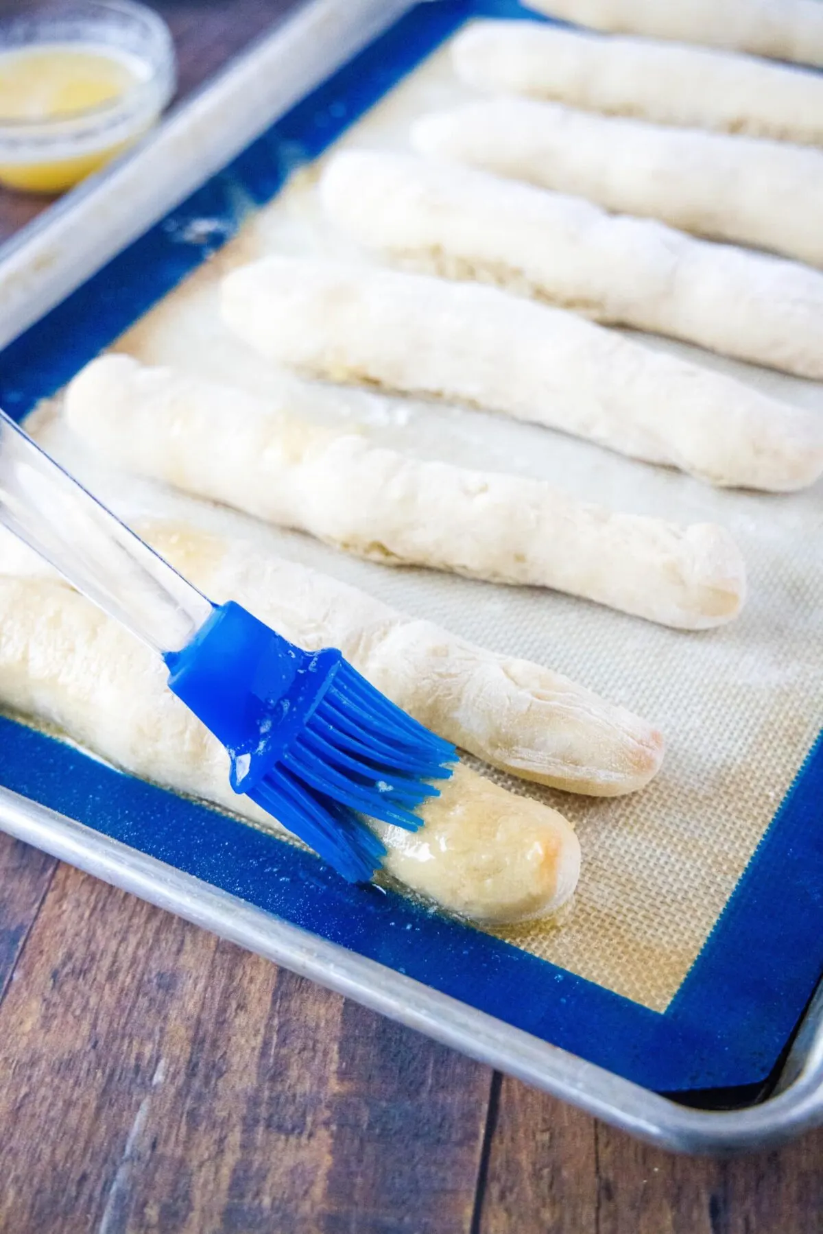 A brush painting butter onto breadsticks on a baking sheet