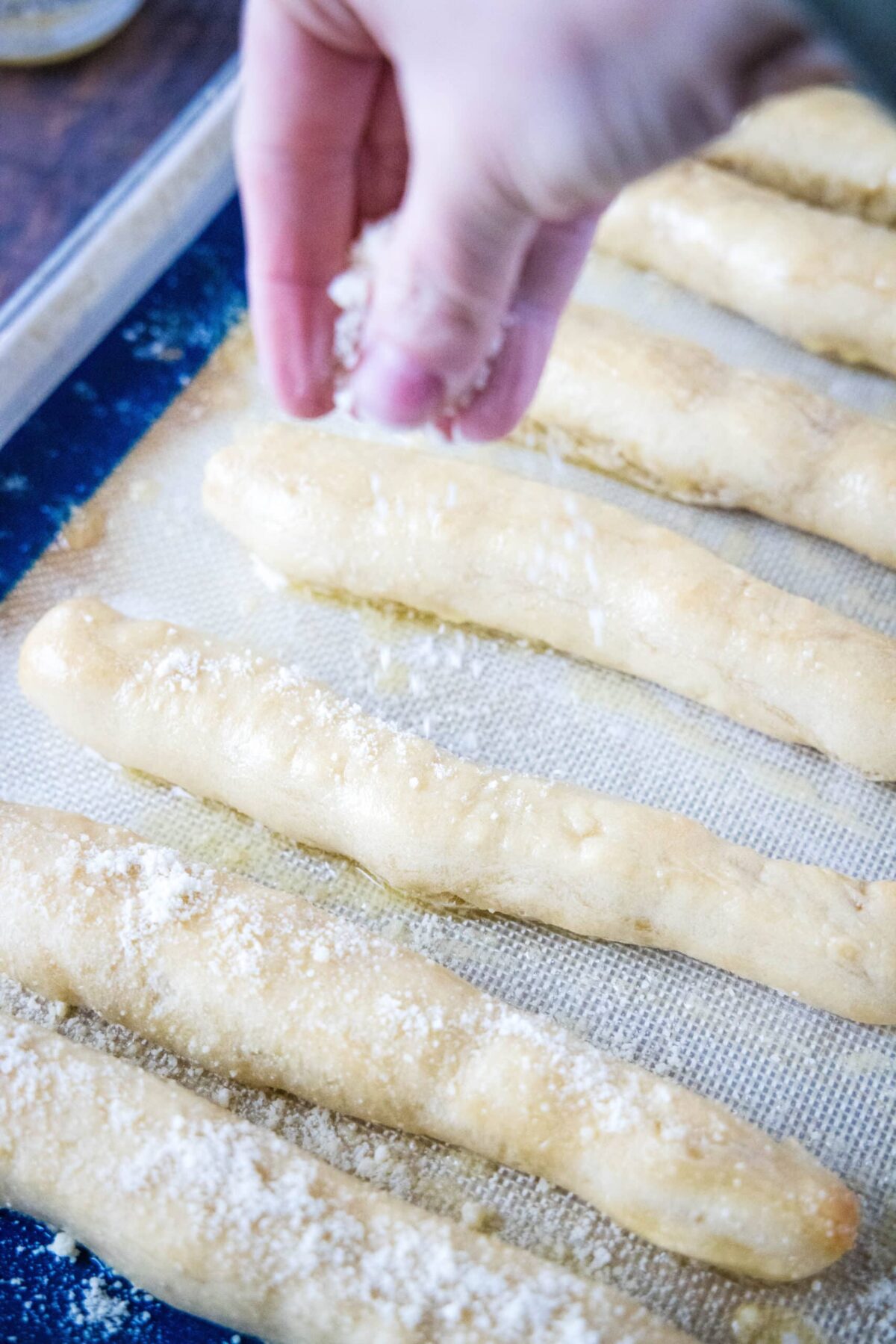 A hand sprinkling parmesan cheese on breadsticks on a baking sheet