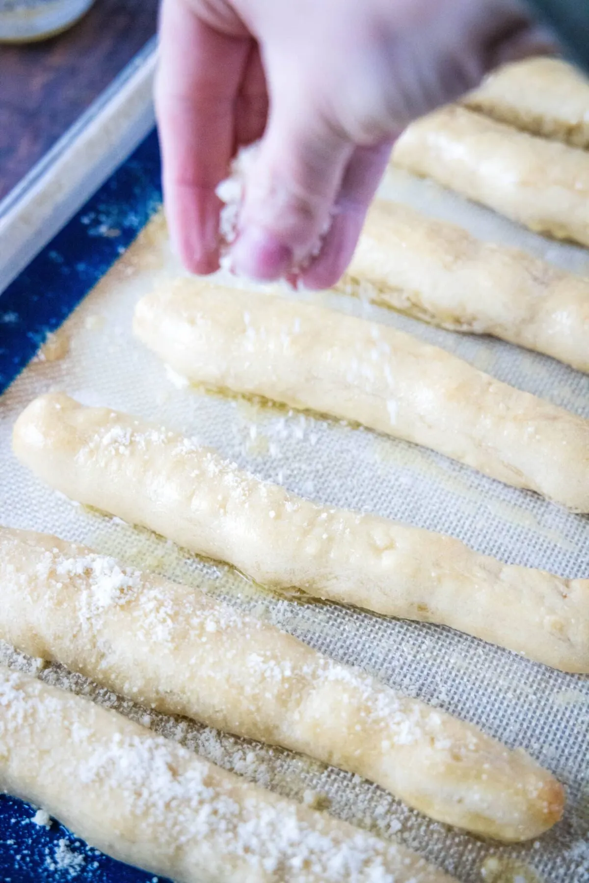 A hand sprinkling parmesan cheese on breadsticks on a baking sheet