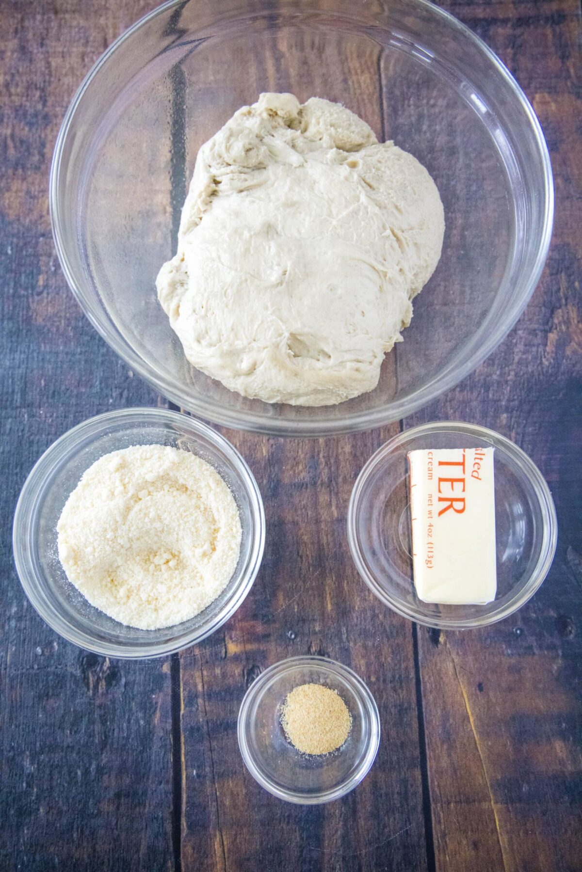Overhead view of the ingredients needed for crazy bread: a bowl of pizza dough, a bowl of parmesan cheese, a bowl of garlic powder, and half a stick of butter