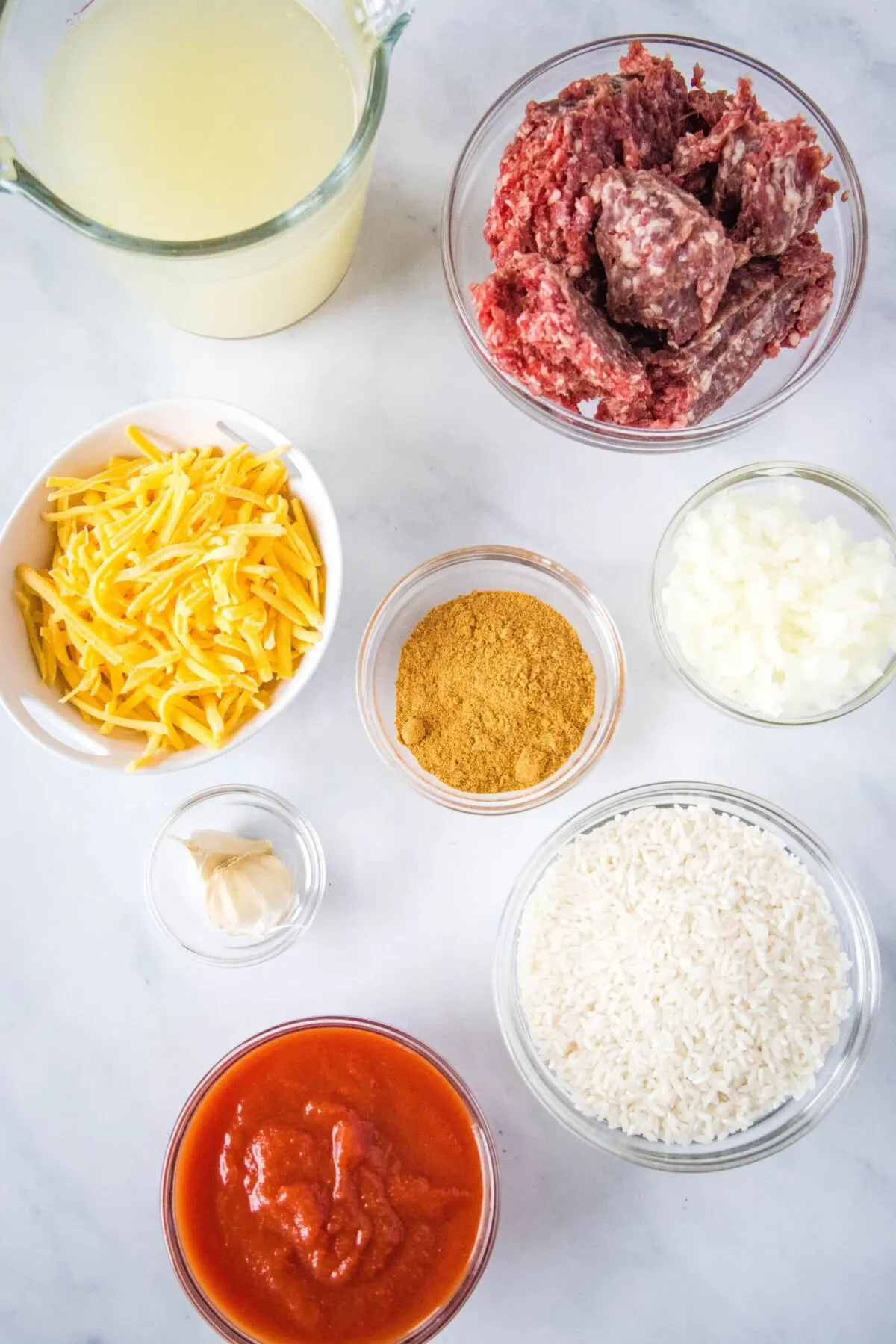 Overhead view of the ingredients needed for taco rice: a bowl of raw ground beef, a bowl of shredded cheddar cheese, a bowl of rice, a bowl of taco seasoning, a bowl of diced onions, a bowl of garlic, a bowl of tomato sauce, and a pitcher of chicken broth