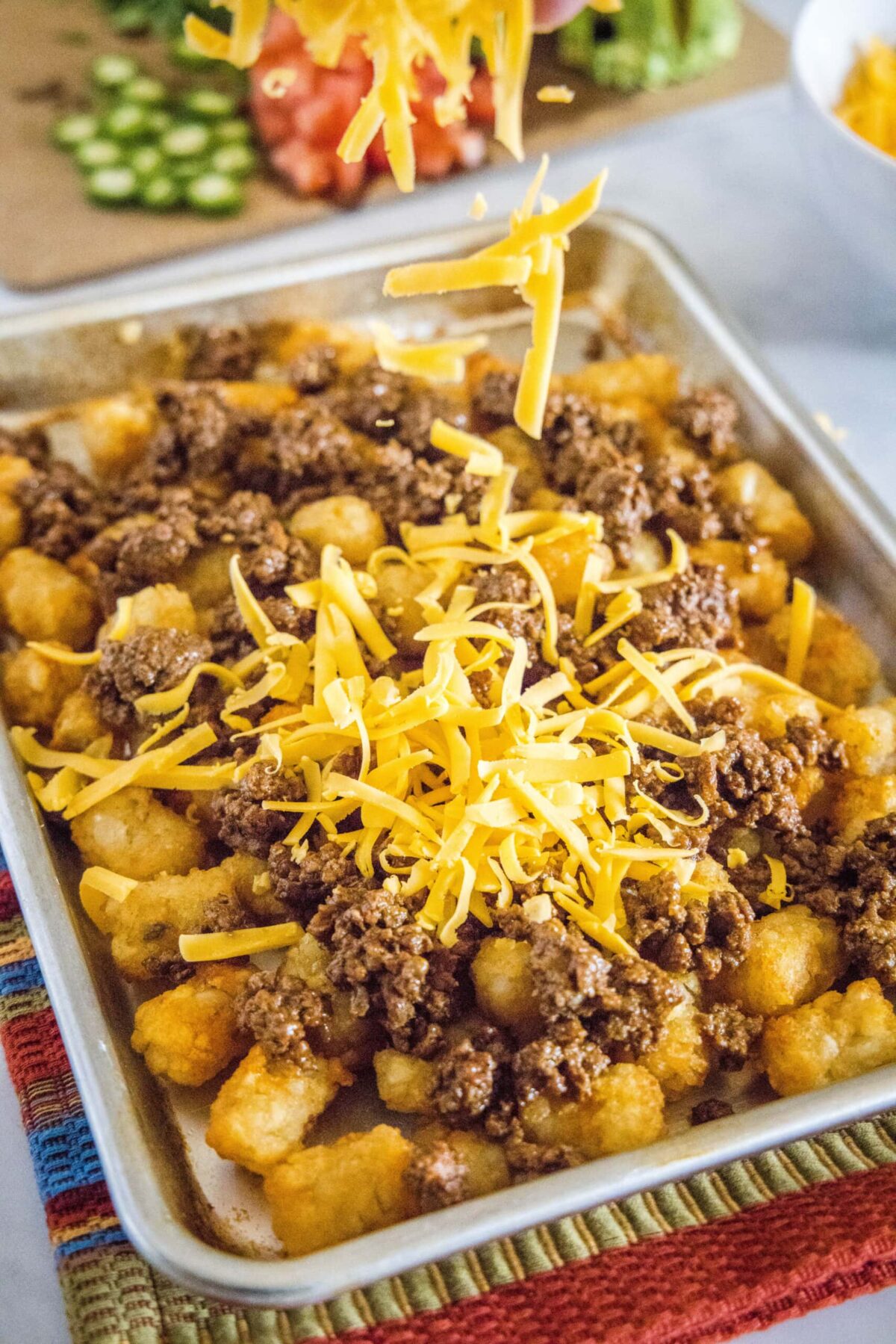 Shredded cheese being dropped onto a baking sheet with tater tots and ground beef