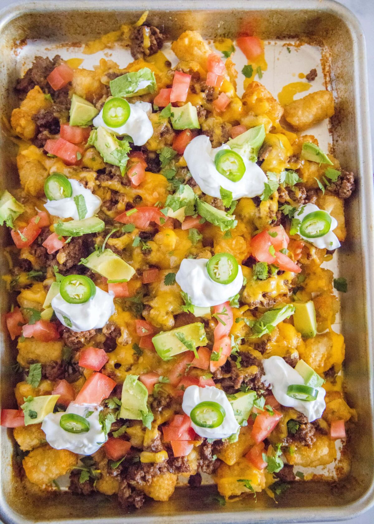 Overhead view of a full baking sheet filled with tater tot nachos, with sour cream, jalapeños, tomatoes, avocado, and cilantro on top