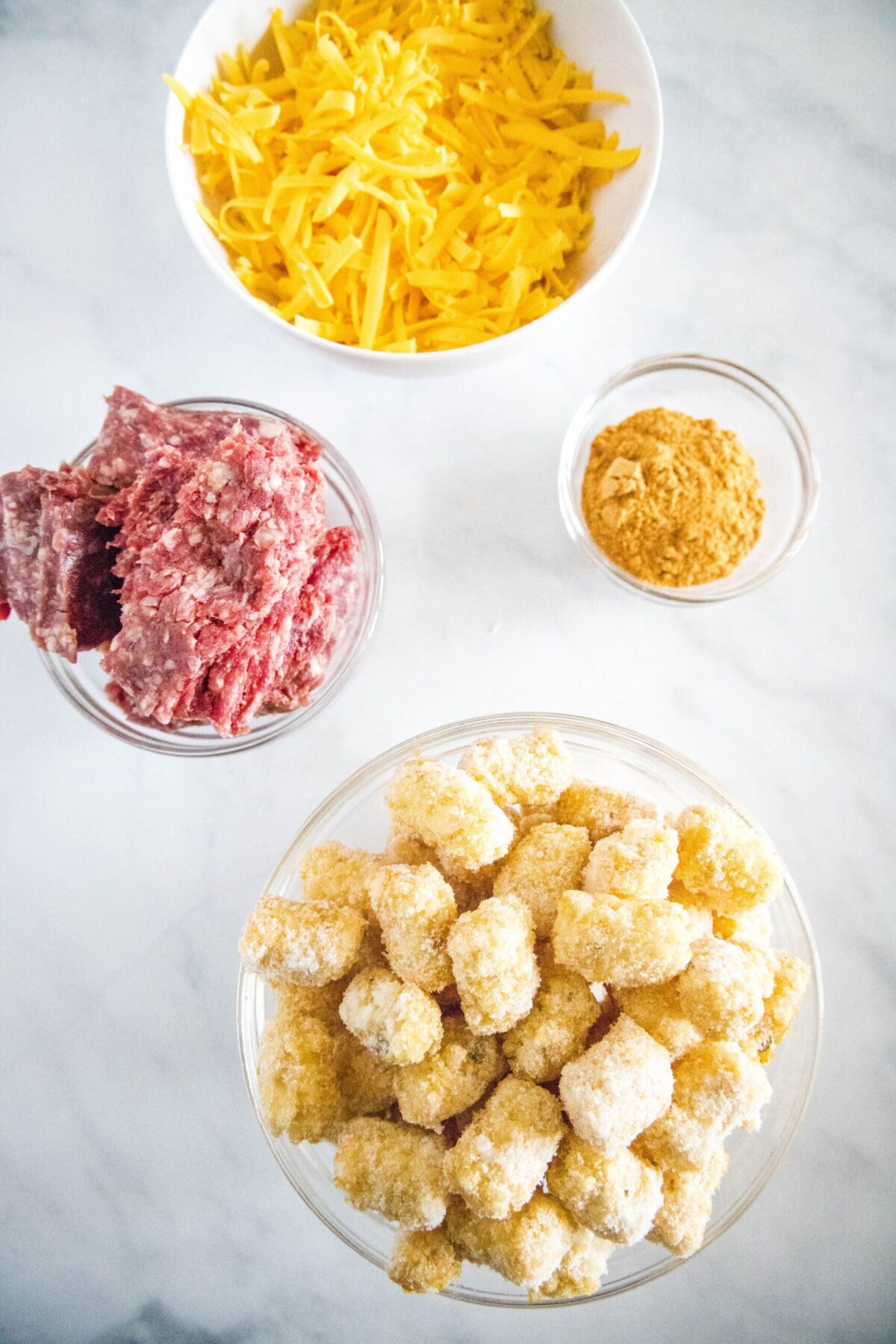Overhead view of the ingredients needed for totchos: a bowl of frozen tater tots, a bowl of raw ground beef, a bowl of shredded cheddar cheese, and a bowl of taco seasoning